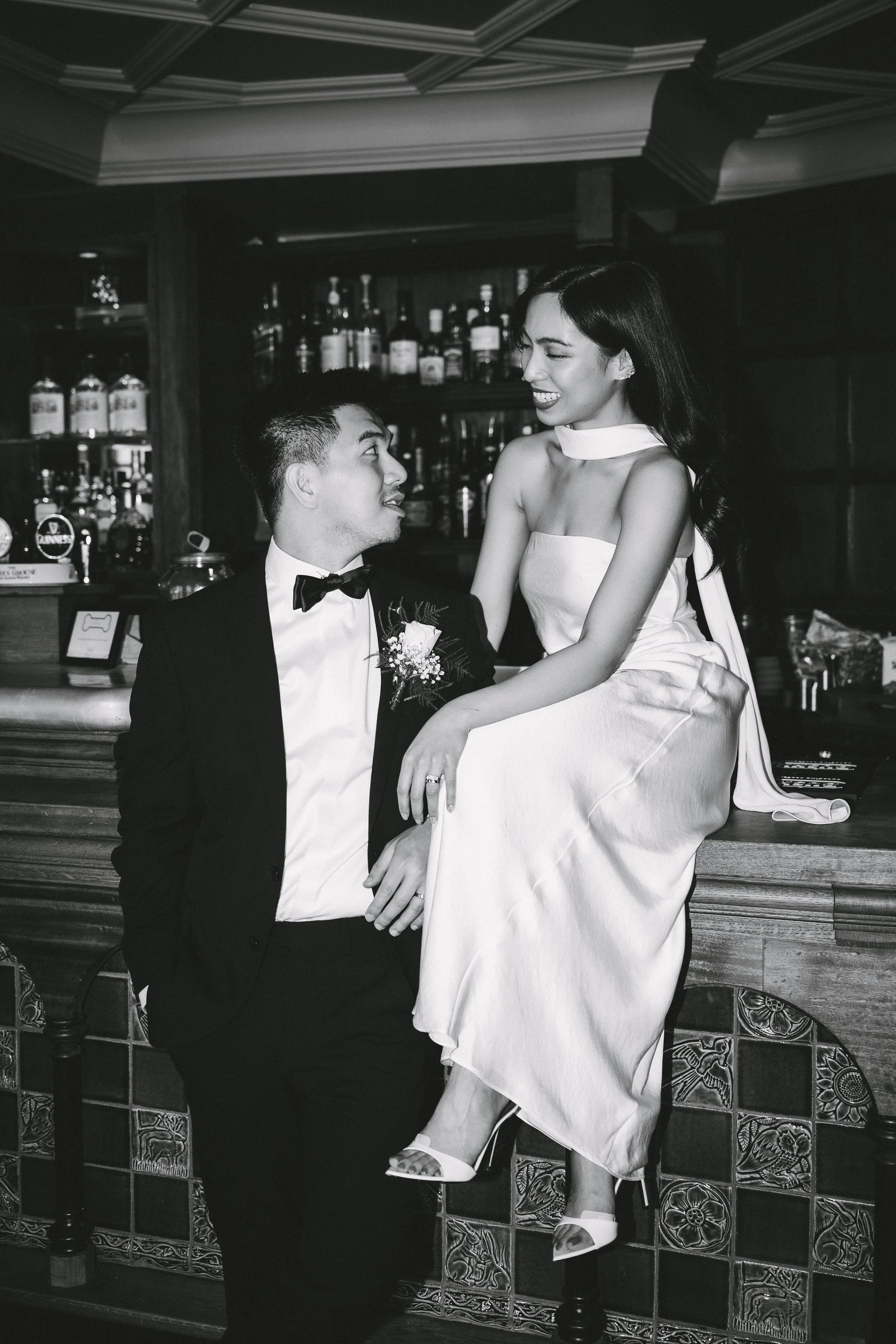 A black and white photo of a couple at a bar, with the woman sitting on the bar counter and the man standing beside her, both dressed in formal wedding attire, smiling at each other.