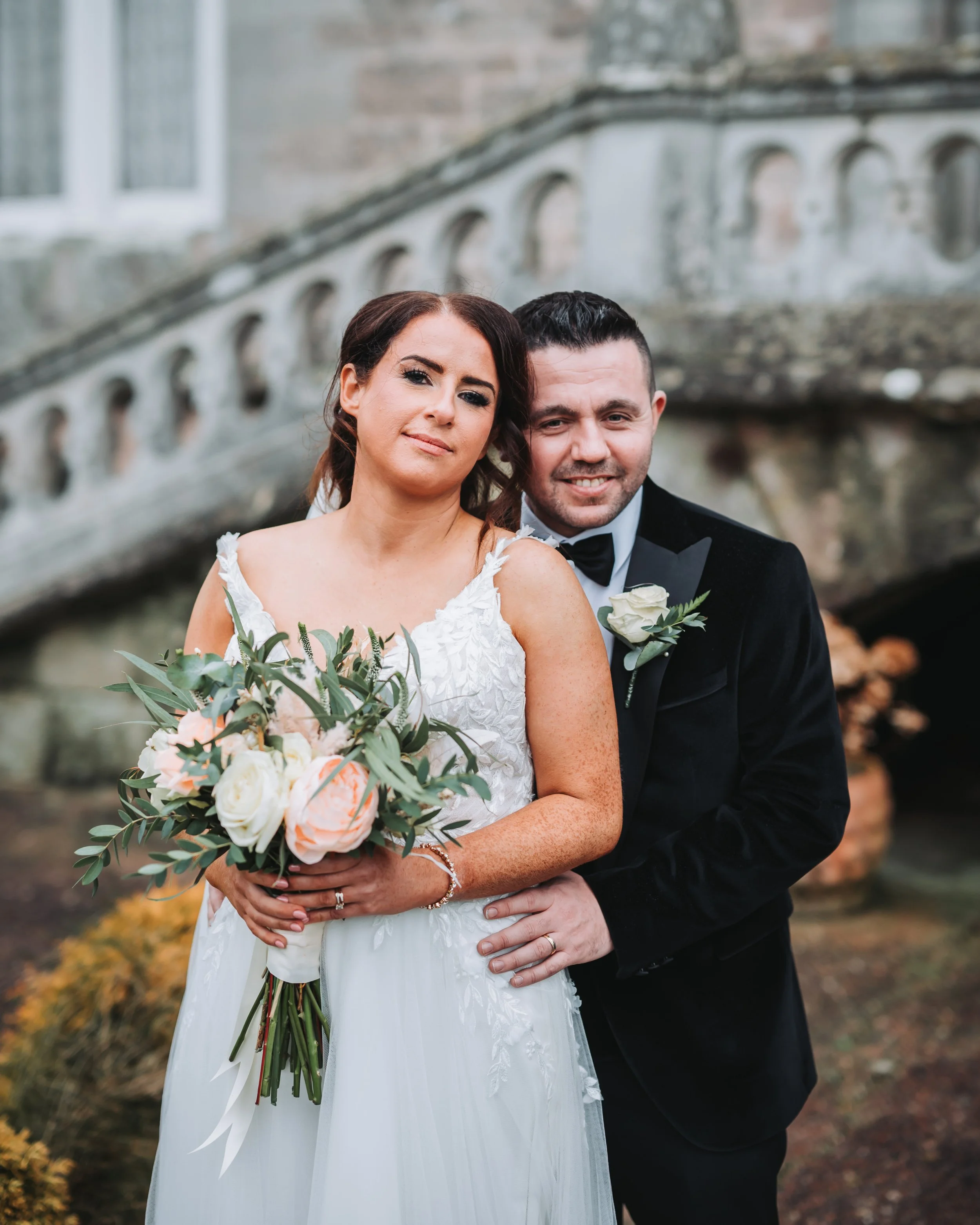 A bride and groom pose for a wedding photo outdoors, with the bride holding a bouquet and the groom in a tuxedo with a white rose boutonniere.