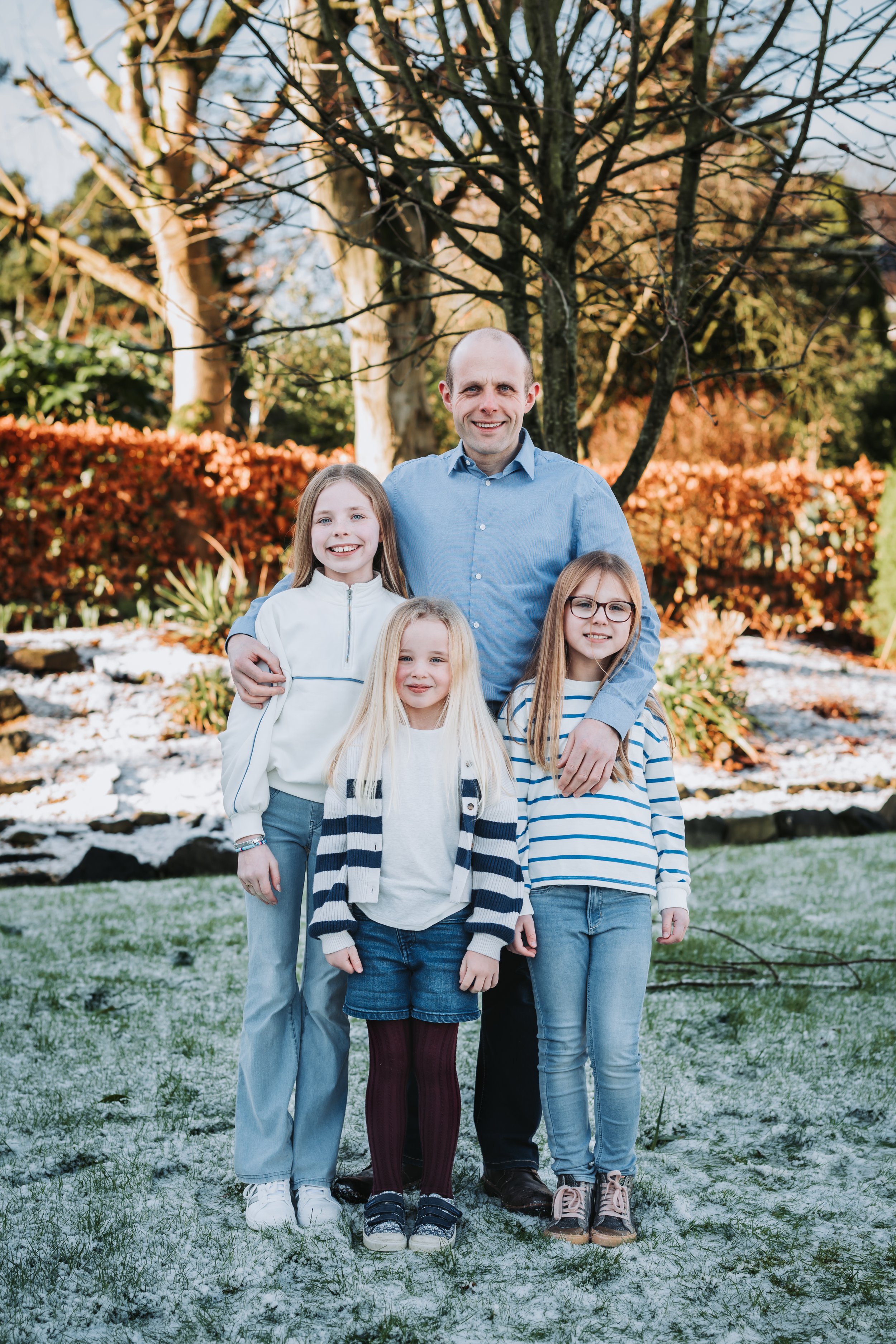 A man and four young girls standing outdoors in a garden with trees and bushes, some snow on the ground, during autumn.