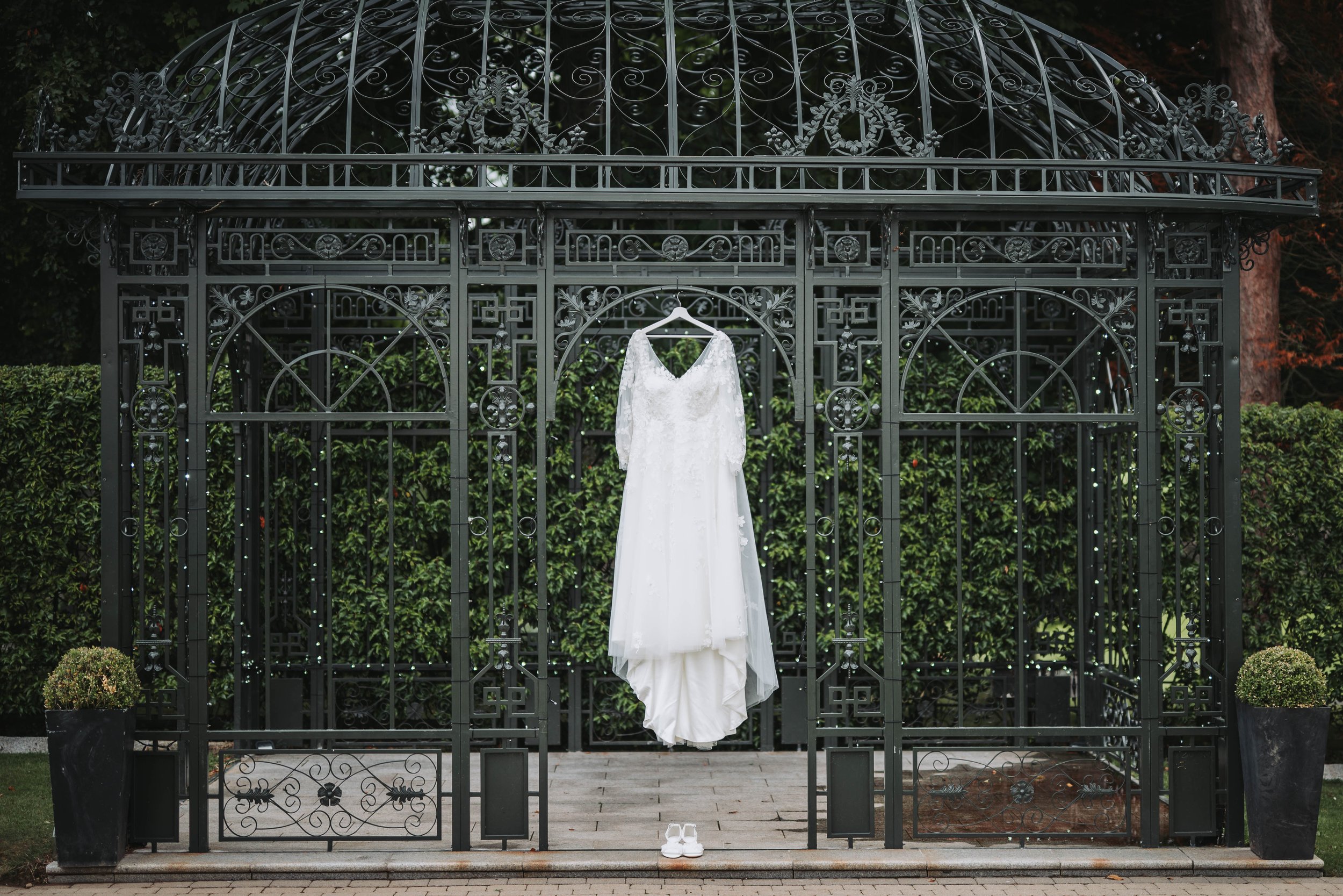 Wedding dress hanging in a black ornate metal gazebo with shoes placed on the ground below.