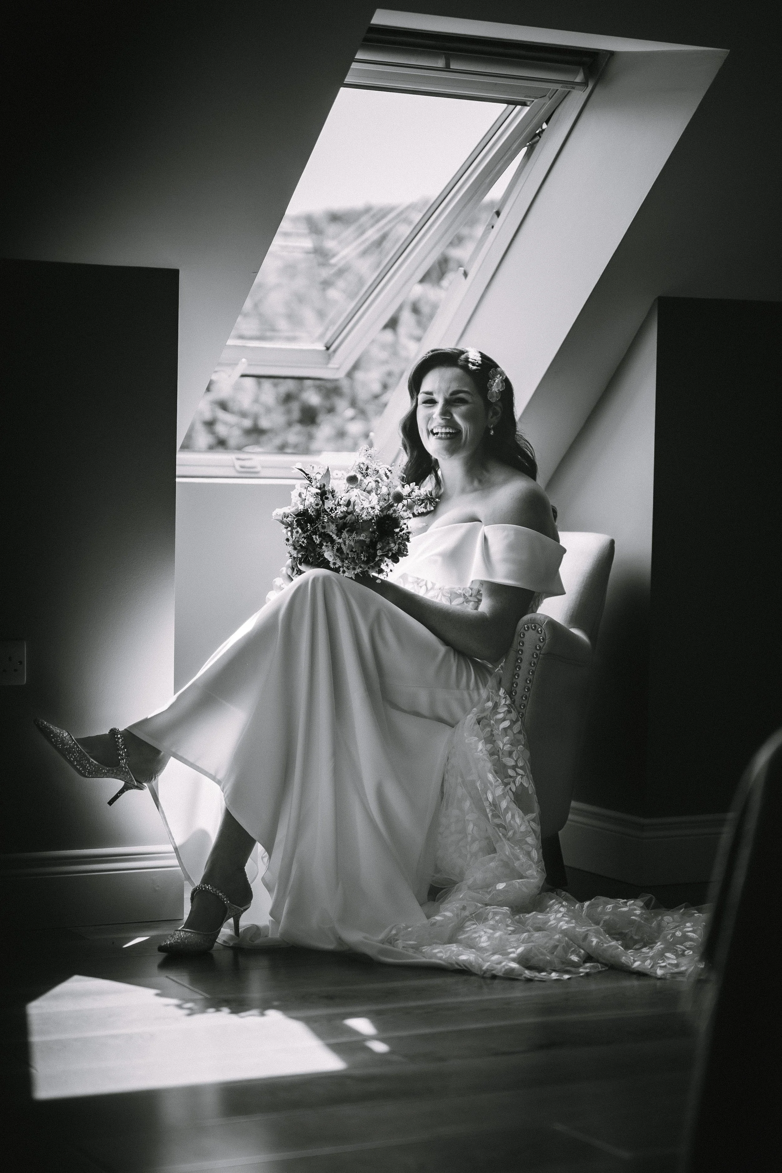 Black and white photo of a woman in a wedding dress sitting on a chair by a slanted window, holding a bouquet of flowers, smiling and laughing.