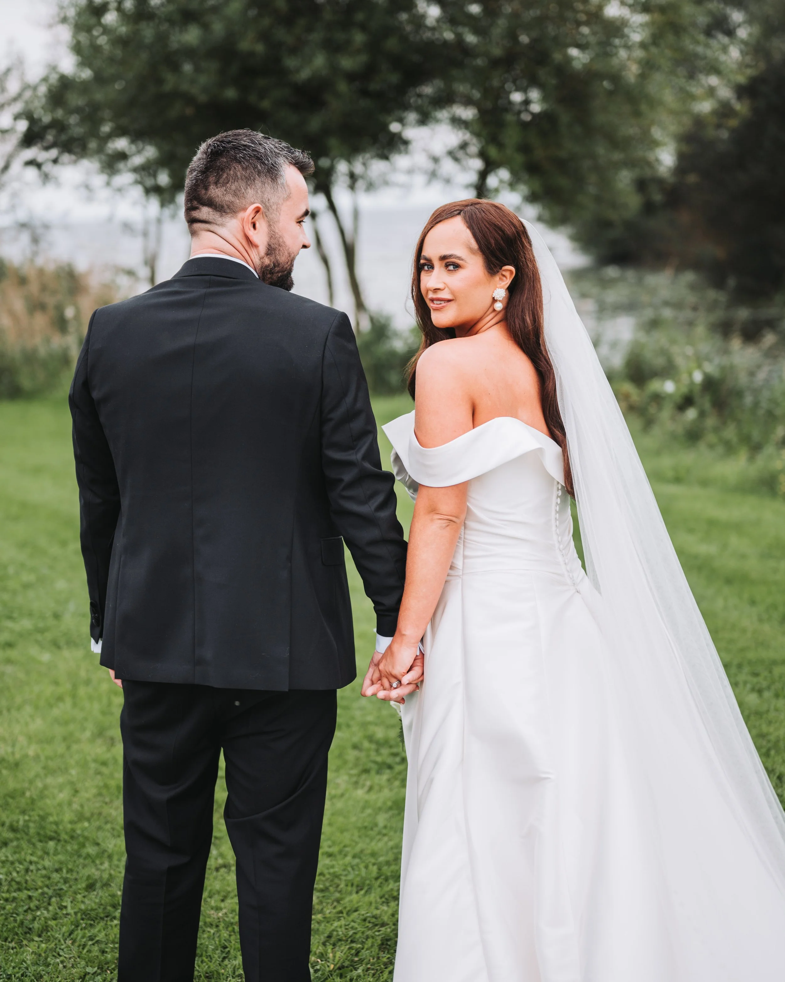 Bride and groom holding hands outdoors, facing away from the camera, with the bride turning her head and smiling. The groom is wearing a black suit, and the bride is in a white wedding gown with off-the-shoulder sleeves, long hair, and earrings. Tree