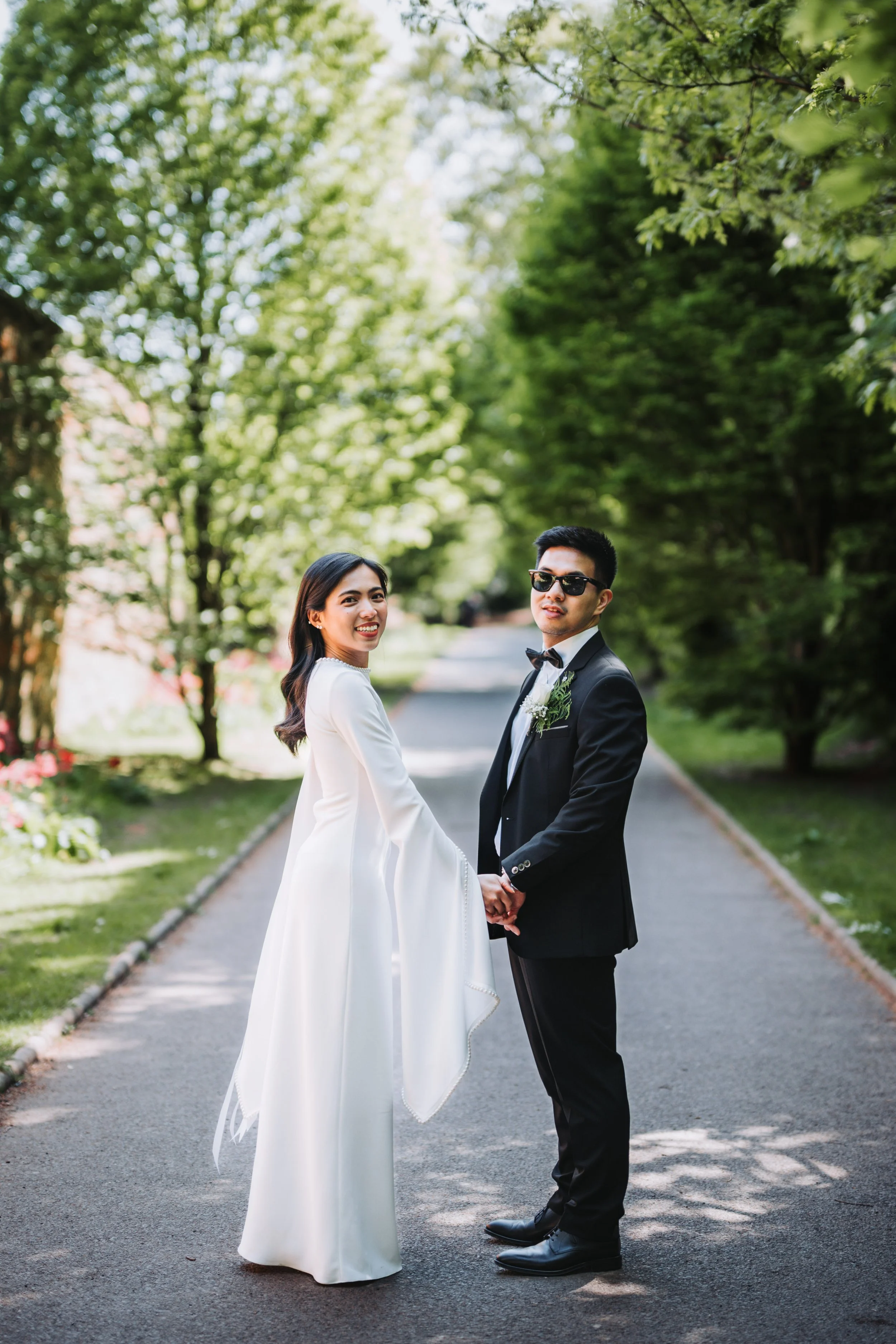 A couple in wedding attire holding hands on a tree-lined street, with the woman wearing a white dress and the man wearing a black tuxedo with sunglasses.