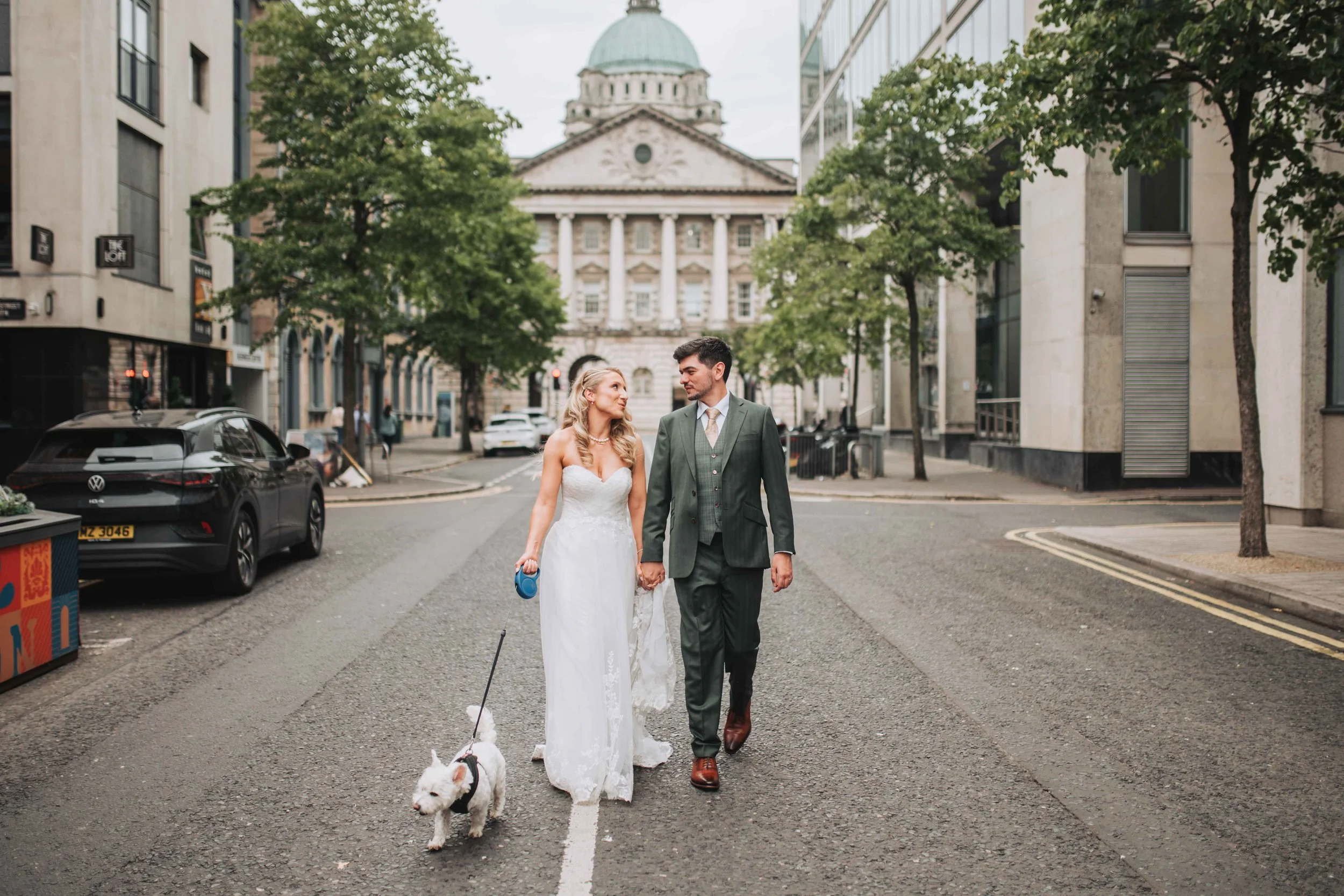 A newlywed couple walking hand in hand on a city street with their dog. The bride in a white wedding dress and the groom in a green suit. They walk past parked cars and modern buildings, with a historic building in the background.