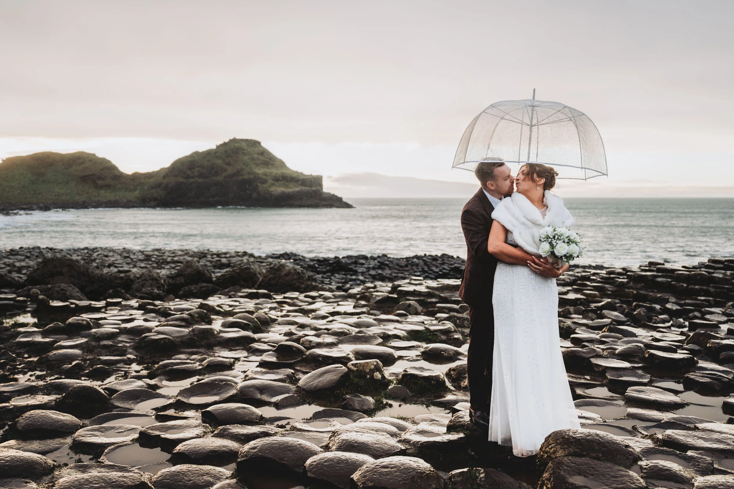 A newlywed couple sharing a kiss under a transparent umbrella on a rocky beach at sunset, with a distant island and calm ocean in the background.