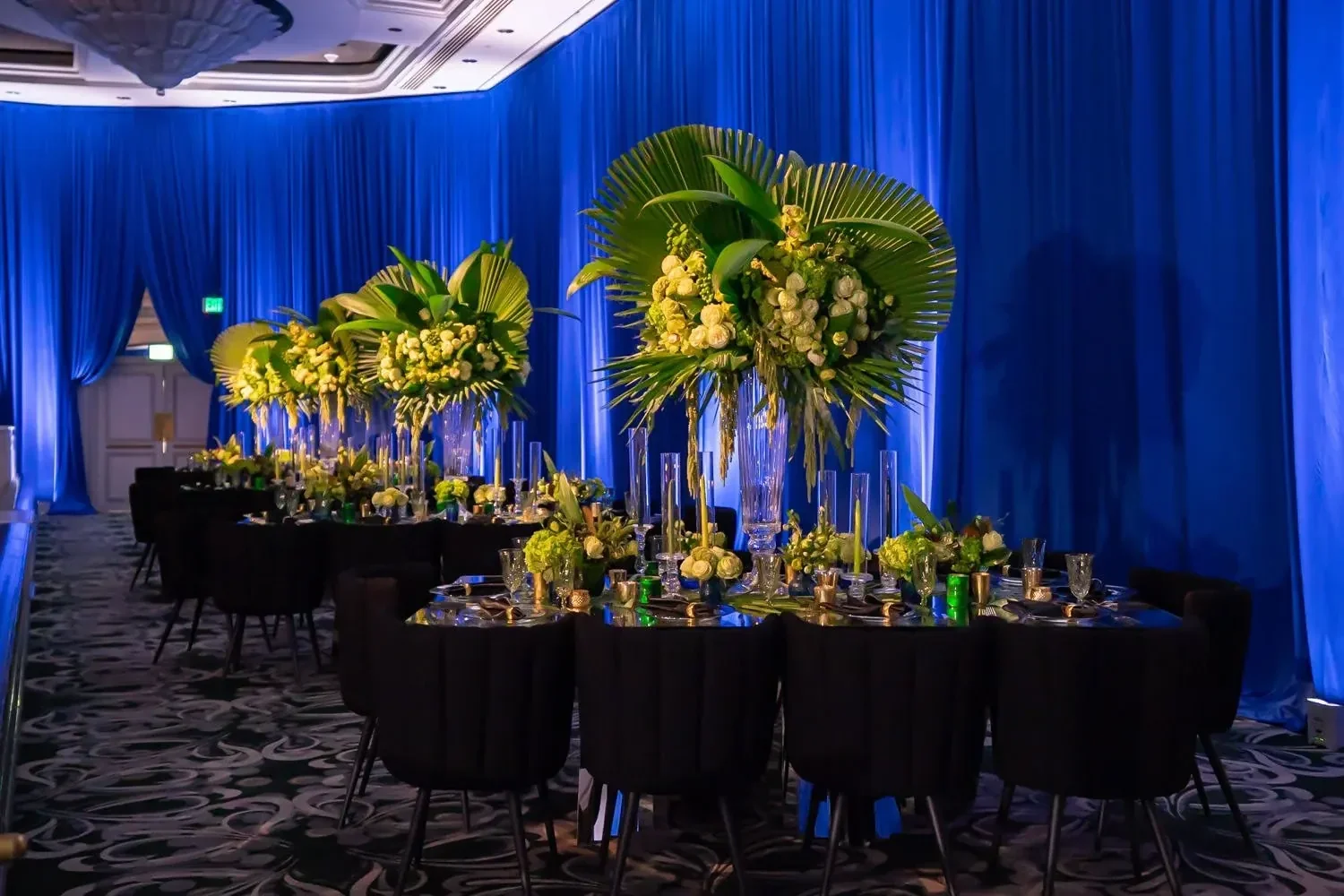 Tropical bar mitzvah reception designed by a party planner, featuring tall palm frond and white floral centerpieces against a royal blue backdrop