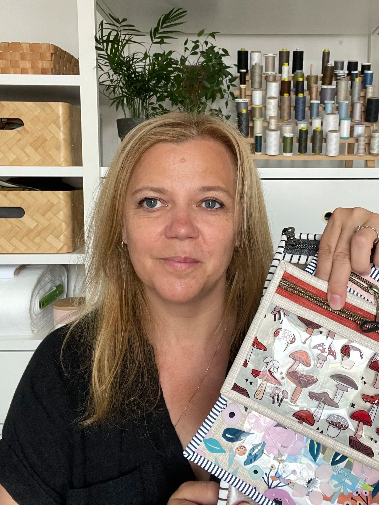 A woman with blonde hair holding a resealable plastic bag with mushroom illustrations, standing in a craft room with thread spools and storage baskets in the background.