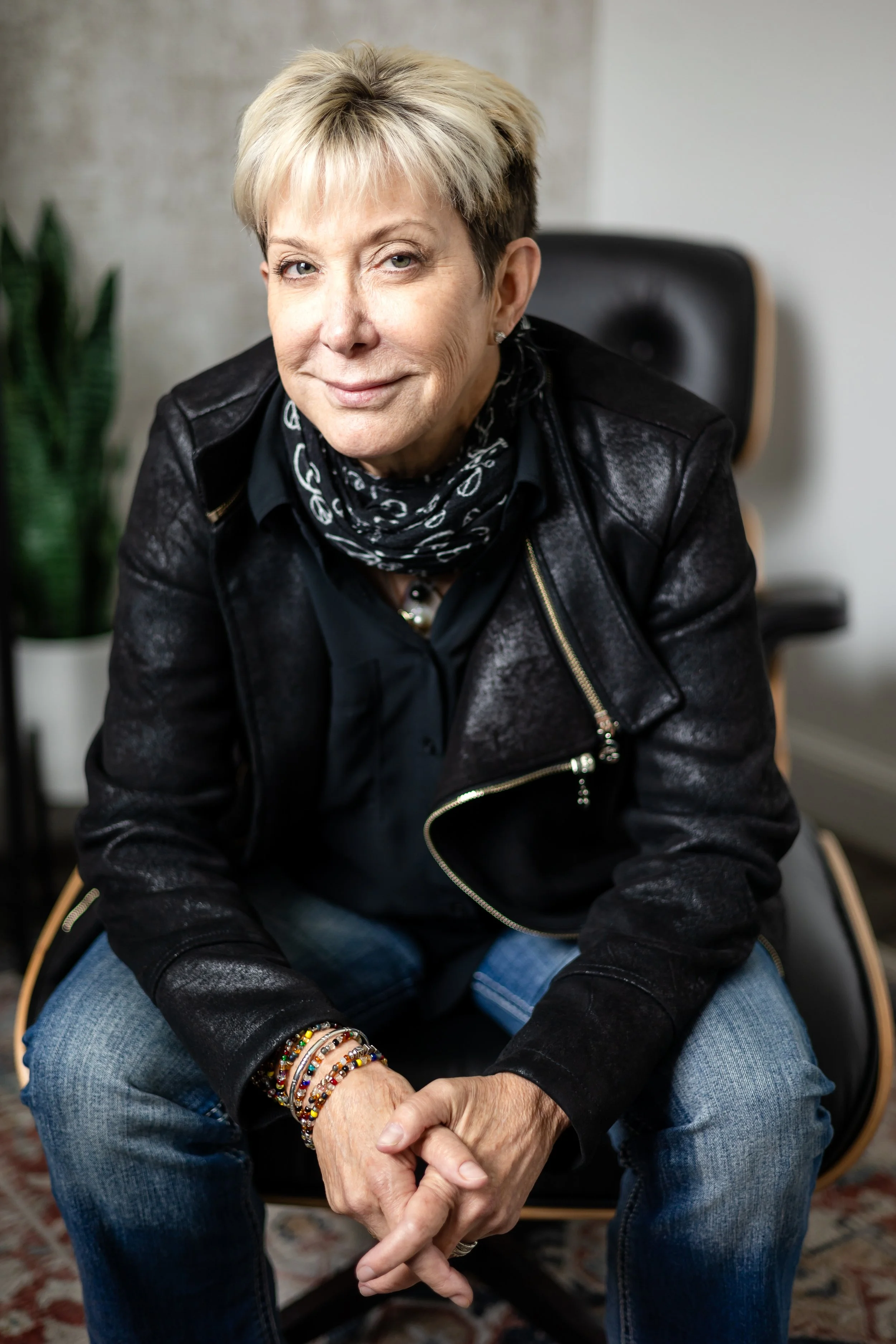 An older woman with short blonde hair, wearing a black leather jacket, black shirt, and blue jeans, sitting on a chair with her hands clasped, smiling slightly, with a plant and a gray wall in the background.