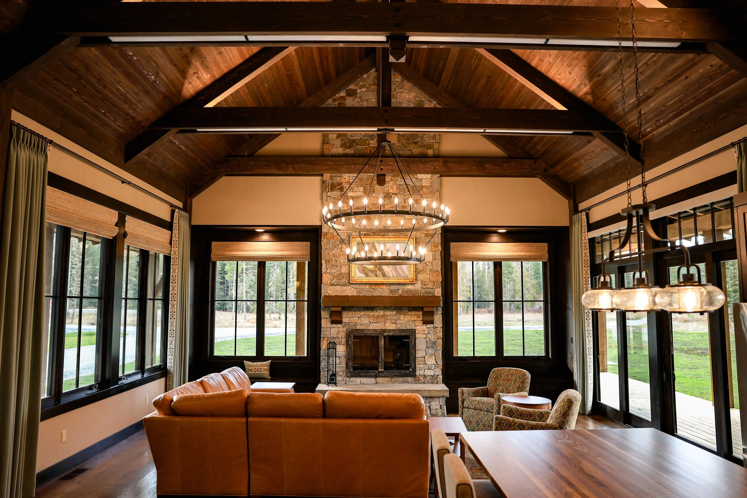 Living room with a brick fireplace, large windows, wooden ceiling with exposed beams, and a chandelier