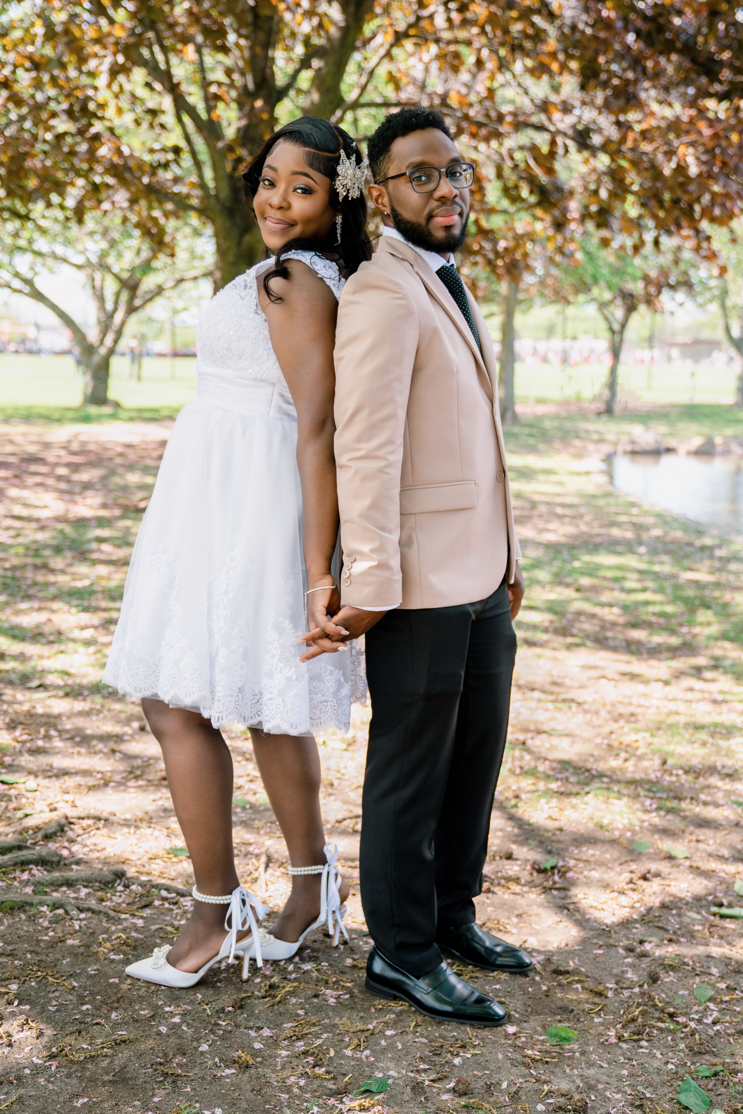A couple holding hands outdoors, standing back to back near a tree, with the woman in a white dress and the man in a beige blazer and black pants, during daytime.