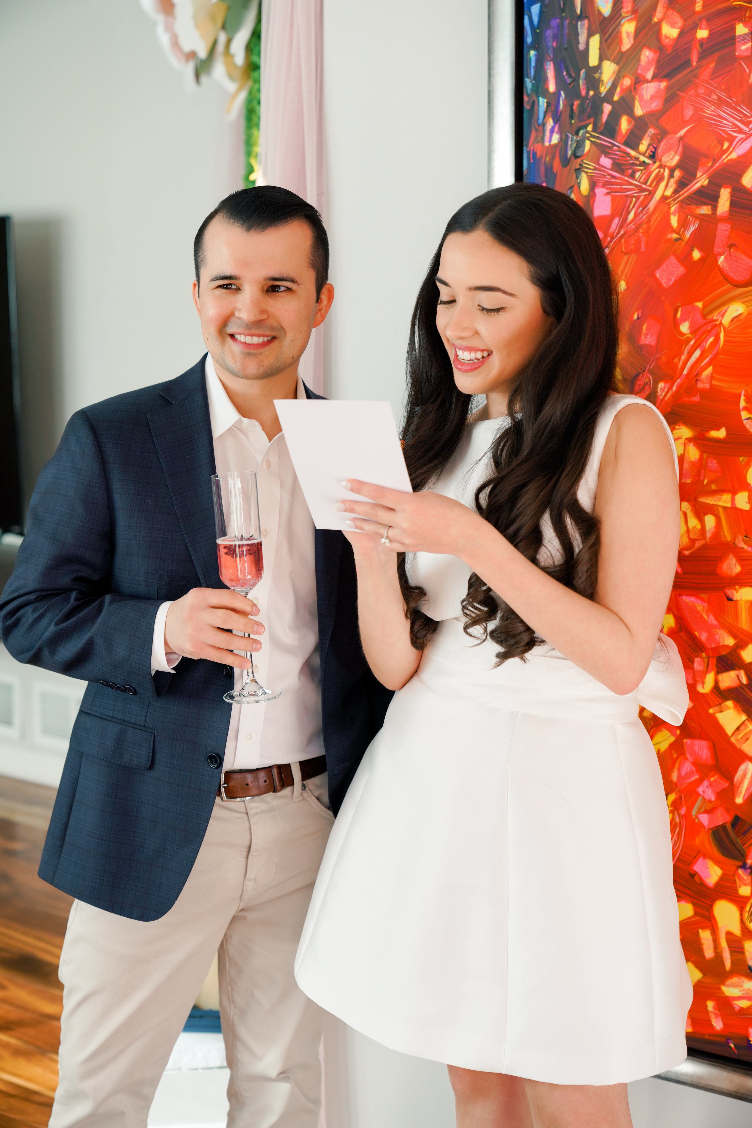 A couple at a celebration, with the man in a navy blazer and beige pants holding a glass of pink beverage, and the woman in a white dress reading a card, standing in front of colorful abstract art.