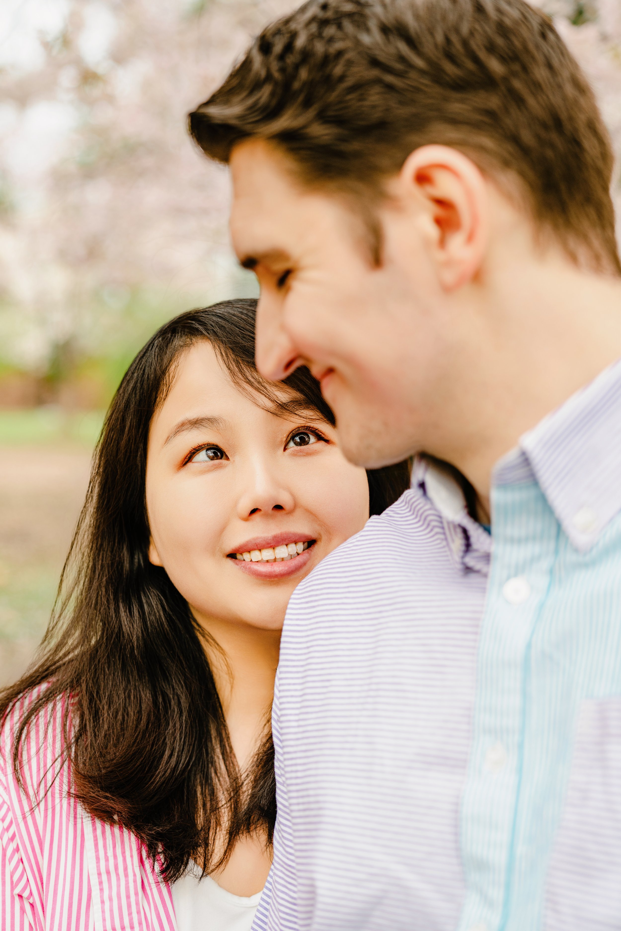 Close-up of a happy couple outdoors, with the woman looking at the man who is slightly smiling, surrounded by a blurred park background.