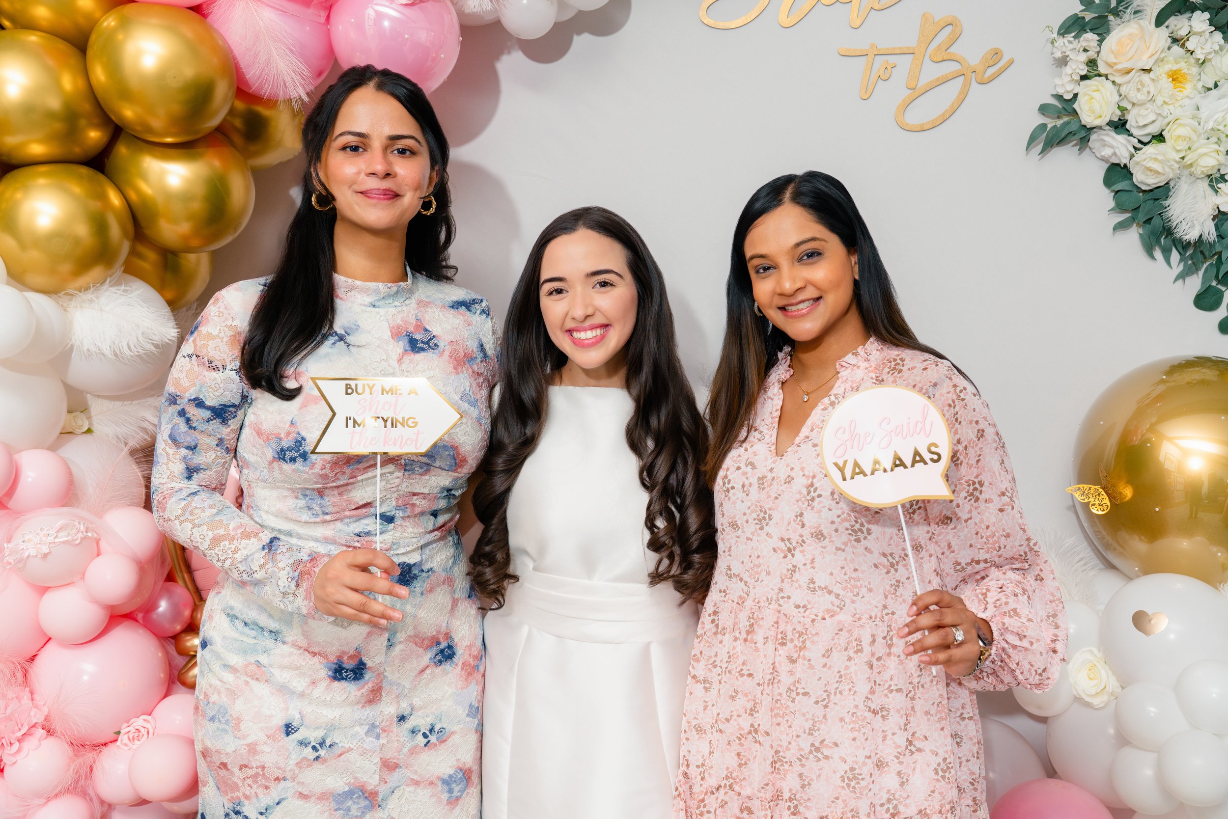 Three women smiling at a celebration with pink, white, and gold balloons, and a floral backdrop. Two women are holding signs with humorous phrases, and another is standing in the middle wearing a white dress.
