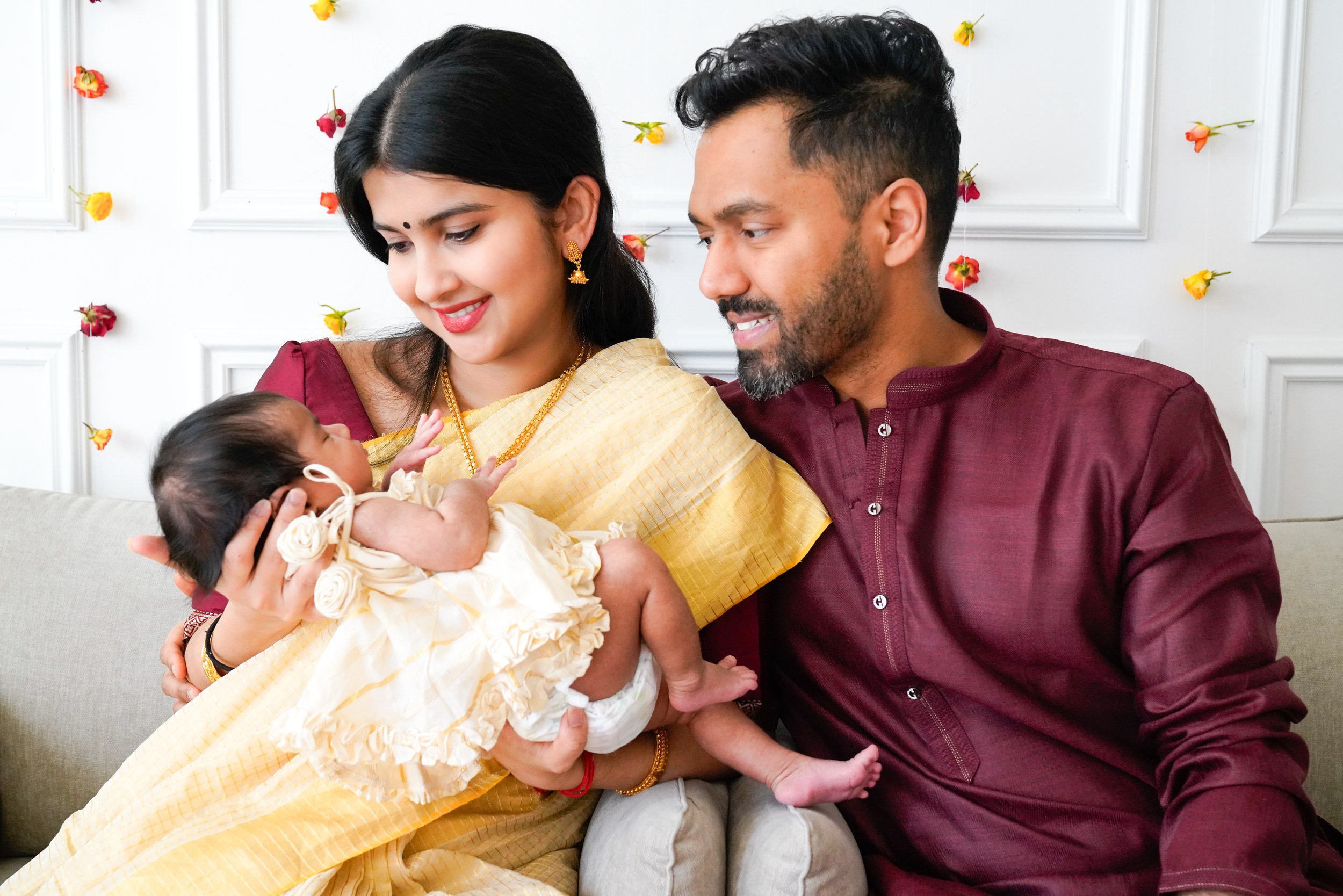 A couple holding a newborn baby, sitting on a sofa with a white wall decorated with small flowers in the background.