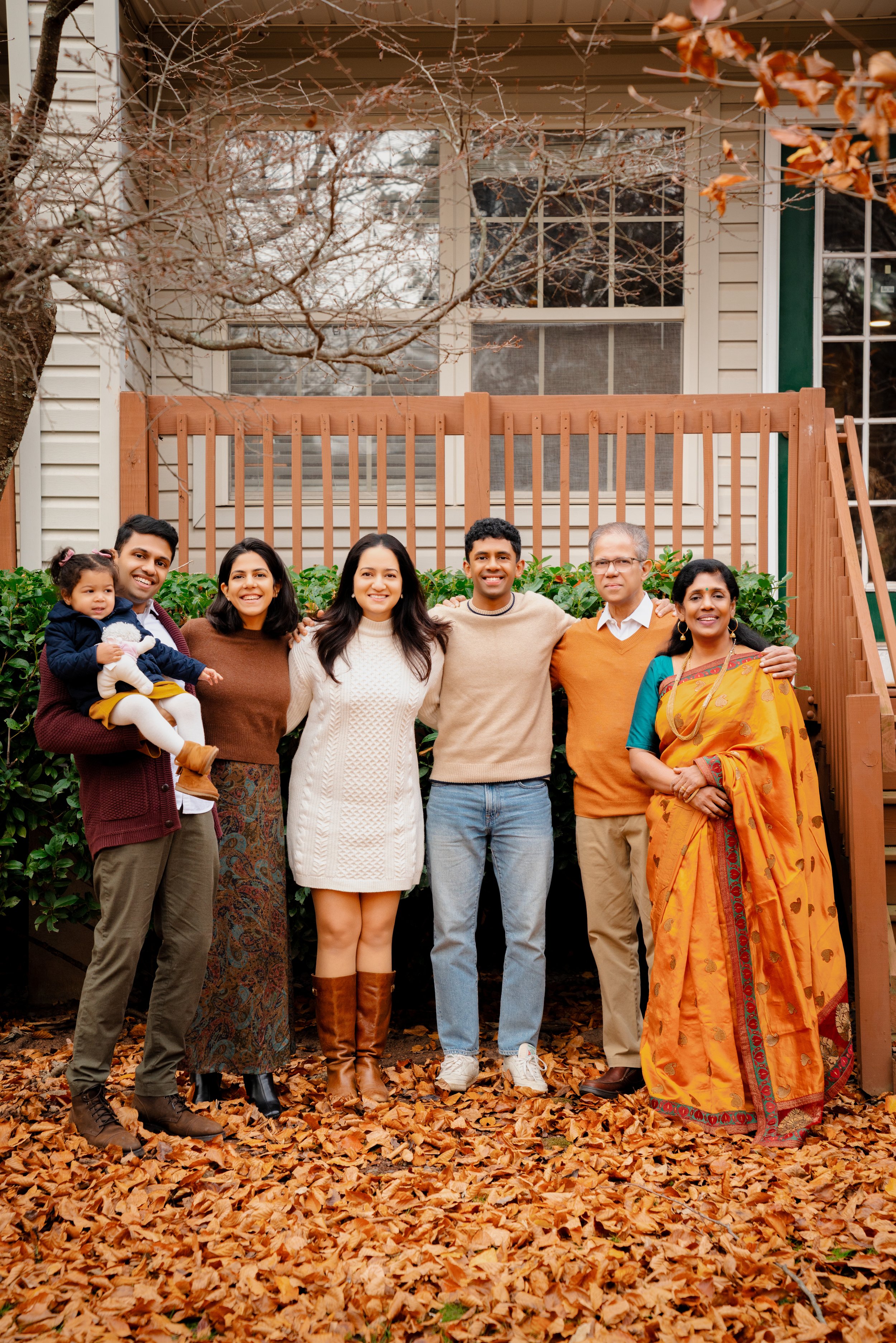 Group of seven diverse people, including a man holding a young girl, standing outdoors on a bed of fallen autumn leaves, smiling for a photo with a house and window in the background.