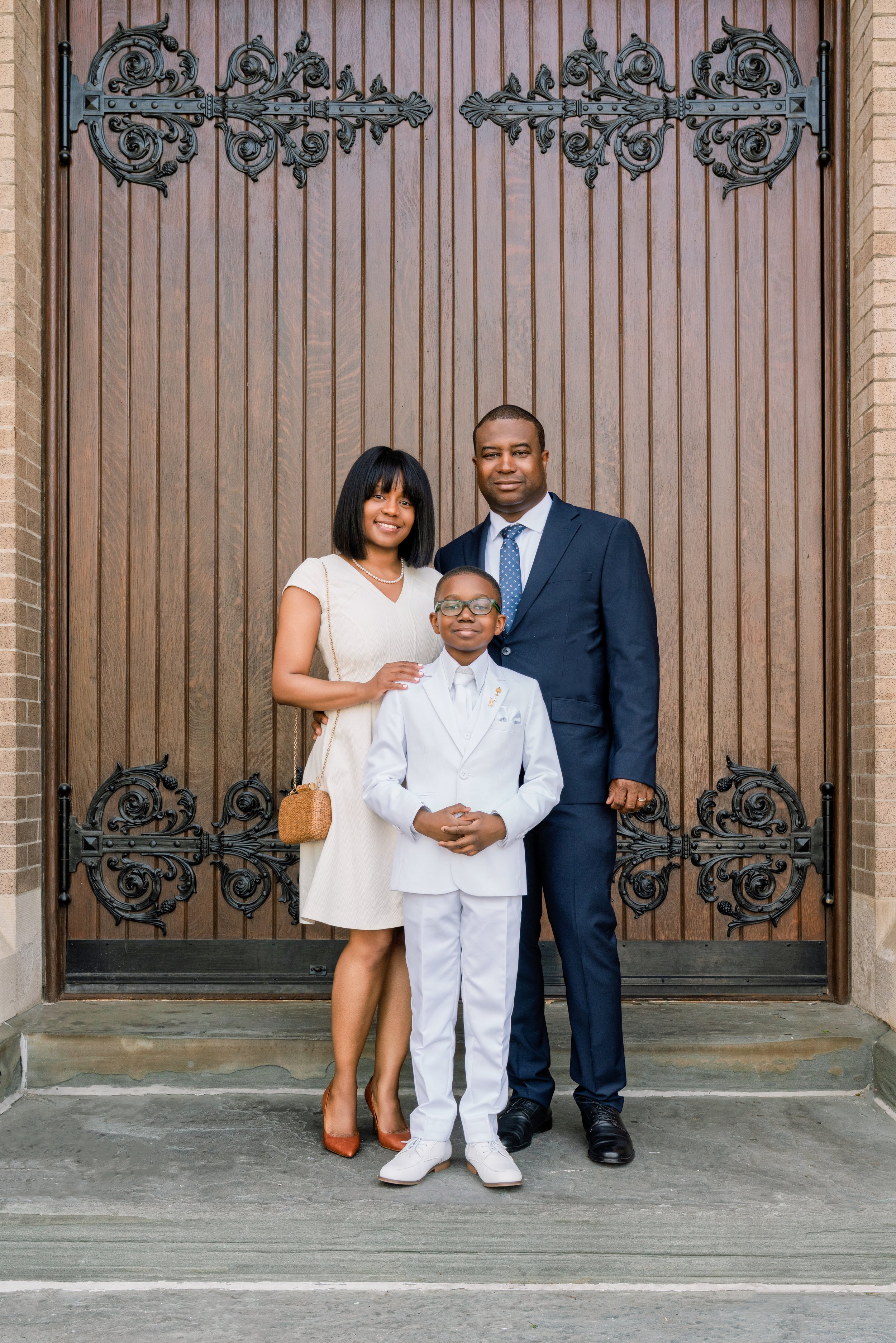 A family of three standing in front of a large wooden door with wrought iron decorative hinges. The mother is wearing a cream dress, the father is in a navy suit, and the young son is dressed in a white suit and glasses. They are smiling, with the mo