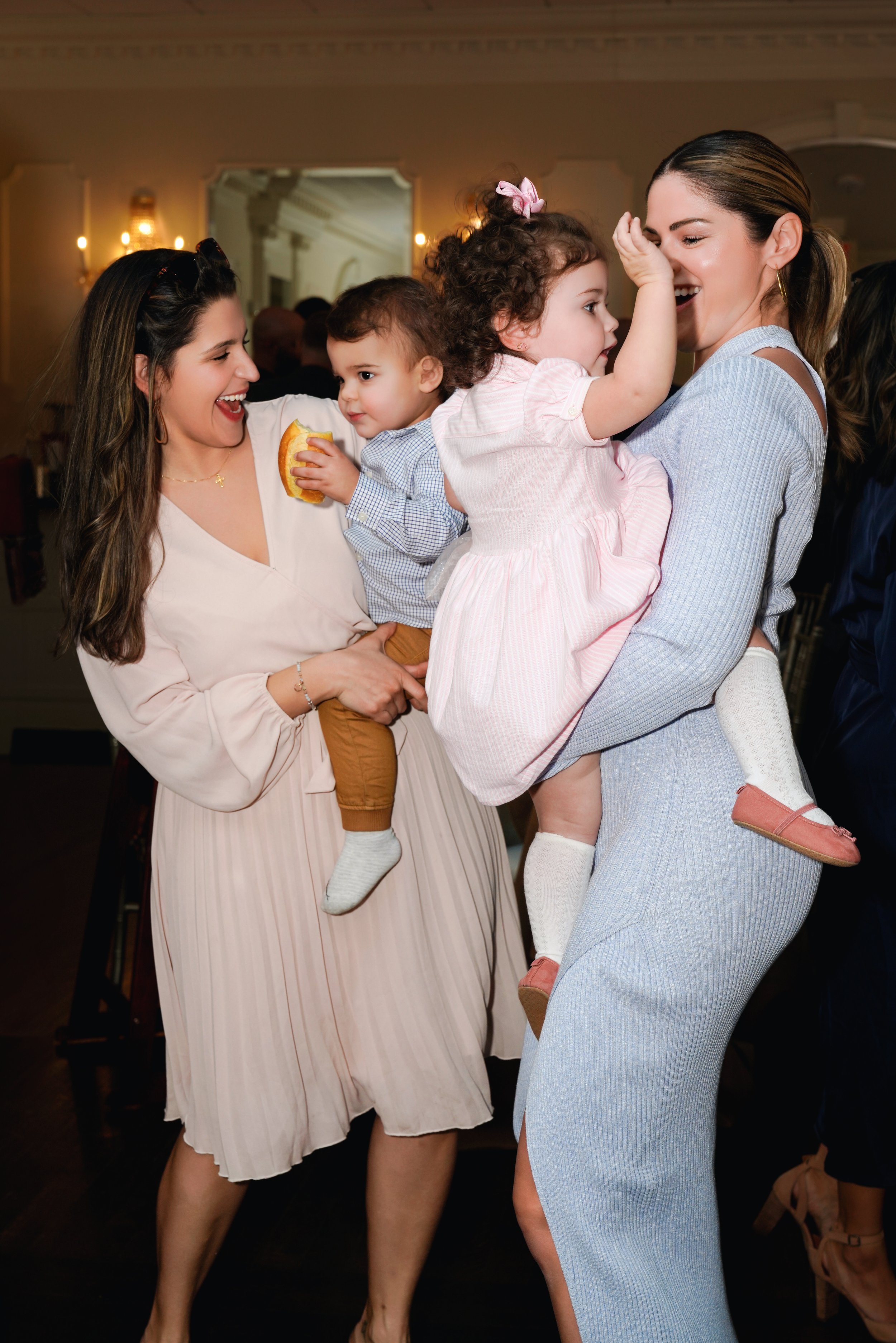Two women holding young children and smiling at each other in an indoor setting, appearing joyful and playful.