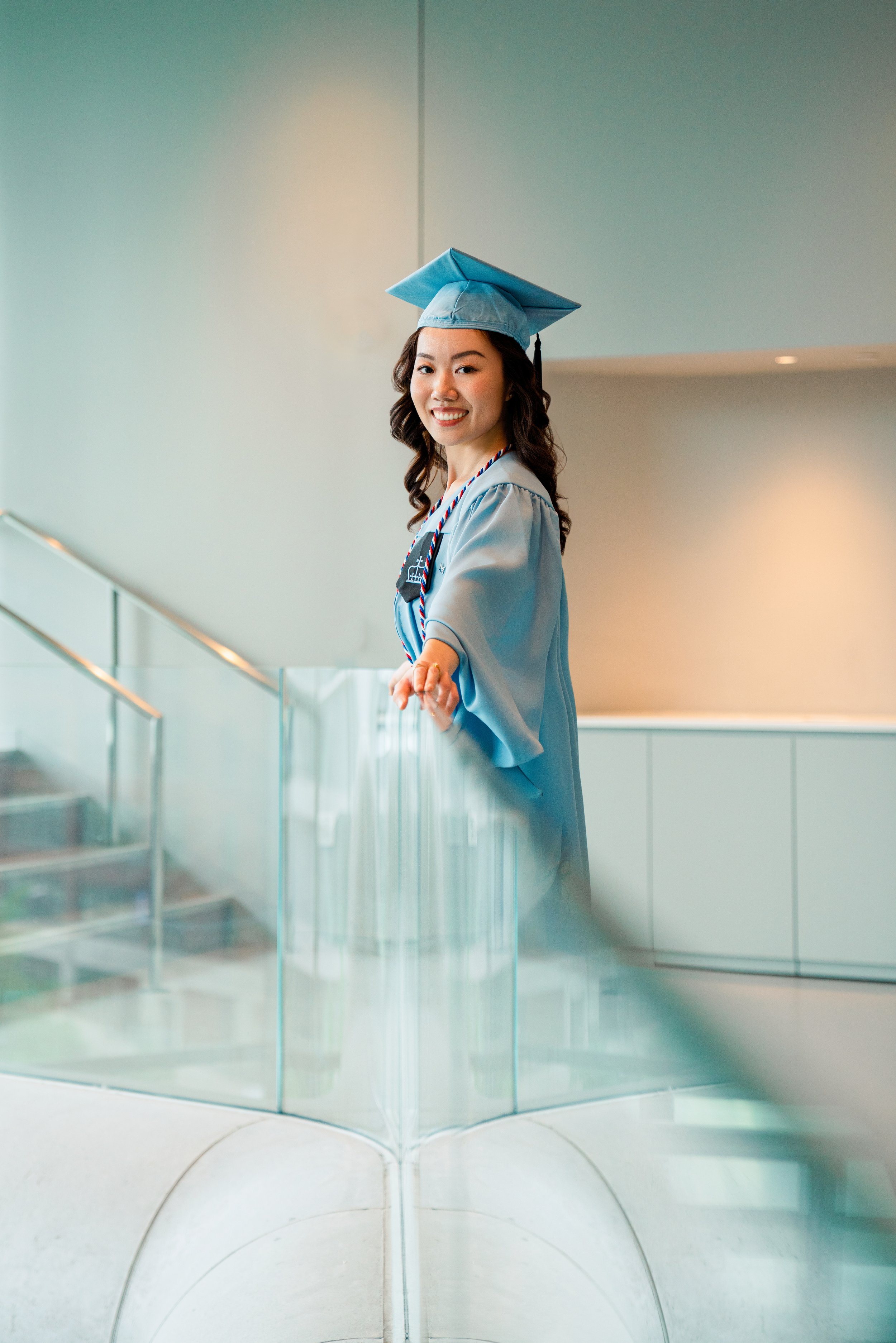 A young woman in a graduation cap and gown smiling and pointing at the camera, standing indoors near a glass railing.