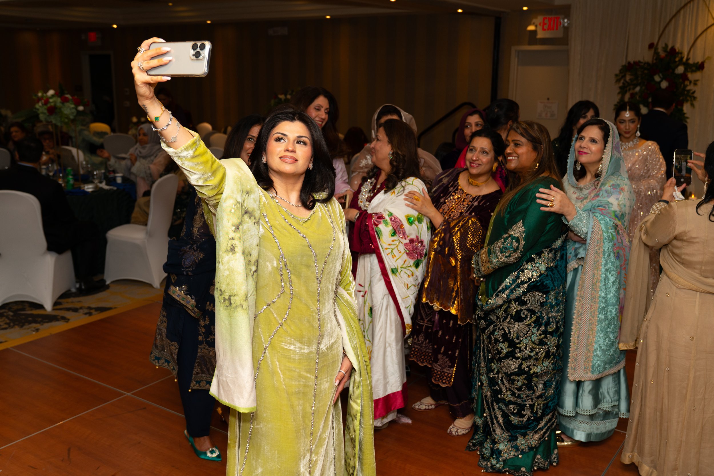 A woman in a yellow traditional dress takes a selfie with a group of women dressed in colorful sarees at a festive event in a banquet hall.