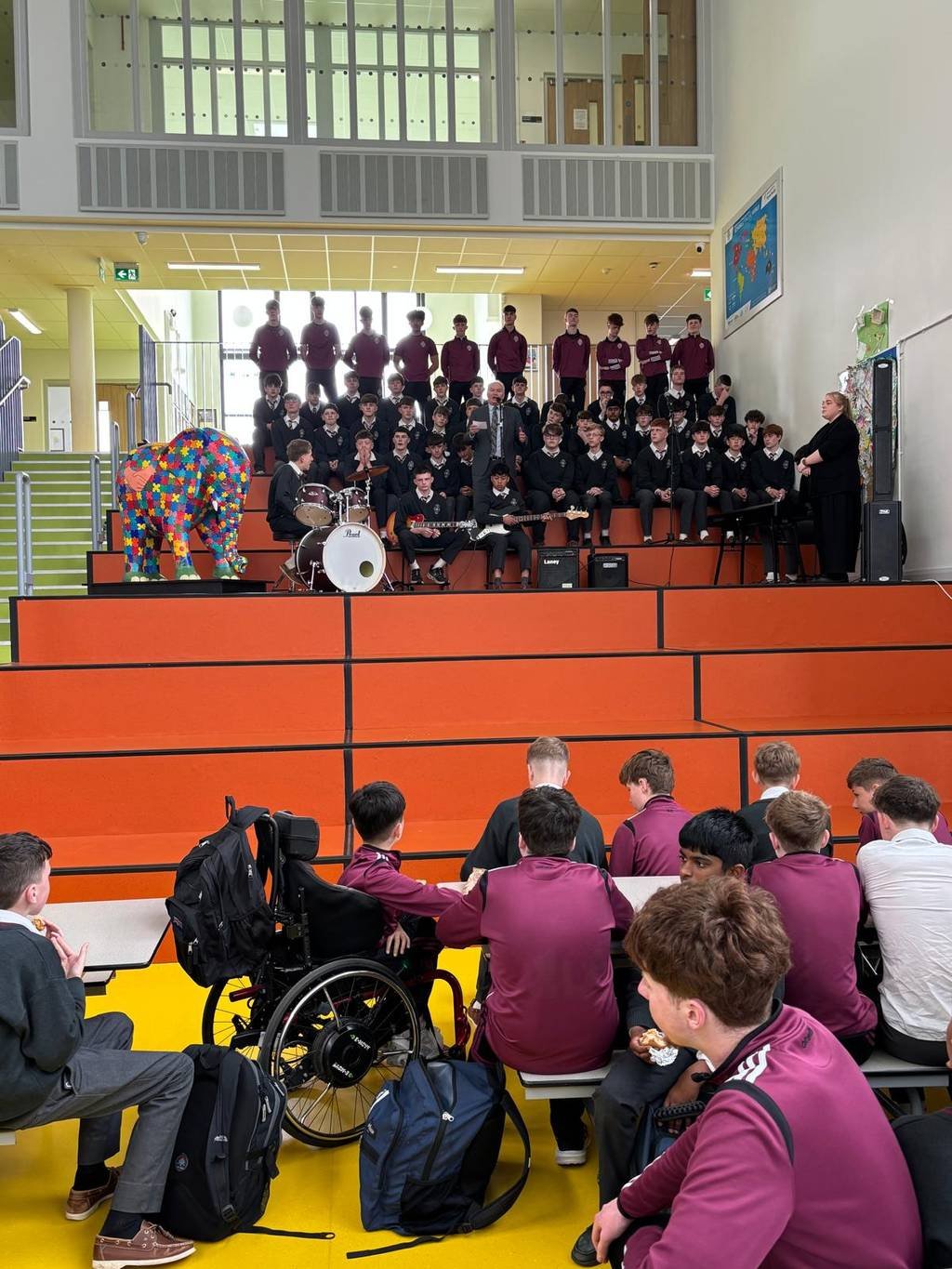 Students gathered on orange tiered seating during a school event, with a speaker at the centre, musicians seated in front, and a brightly painted Elephant in the Room sculpture displayed to the left.