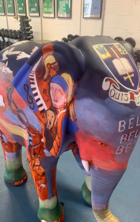 A life-sized elephant sculpture displayed indoors in a gym setting, painted in vibrant red, blue, and green tones. The artwork features detailed illustrations including a piano keyboard, musical notes, a portrait of a child, symbolic imagery, and a h