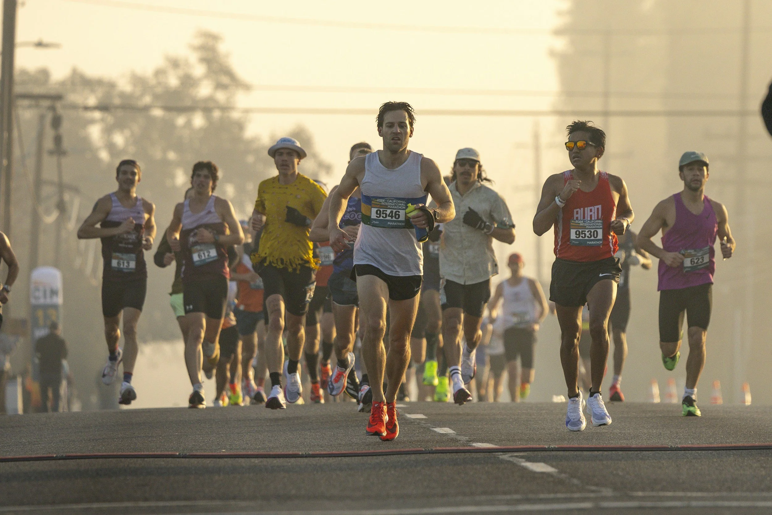 A group of marathon runners participating in a race during sunrise or sunset, running on a paved road with traffic lights and power lines visible in the background.