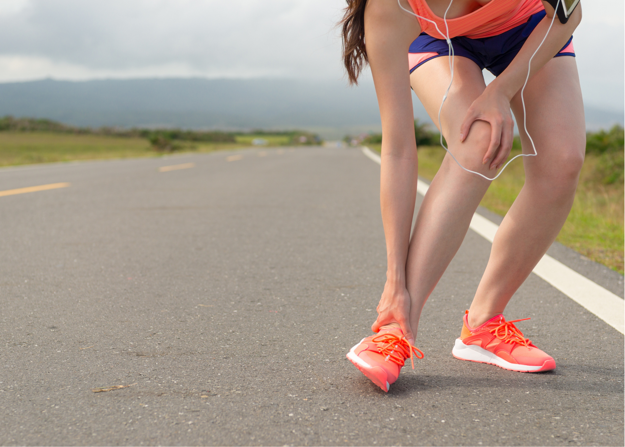 A woman in athletic clothing and bright pink running shoes is bent over on the side of a rural road, holding her knee, appearing to experience discomfort or injury.