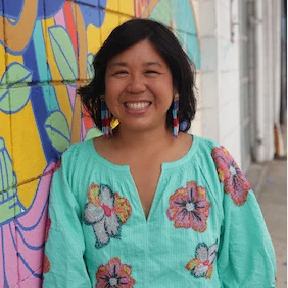 Sharon NG an asian woman smiling in a turqoise blouse with flowers printed on the blouse. Leaning against a colorful mural wall.