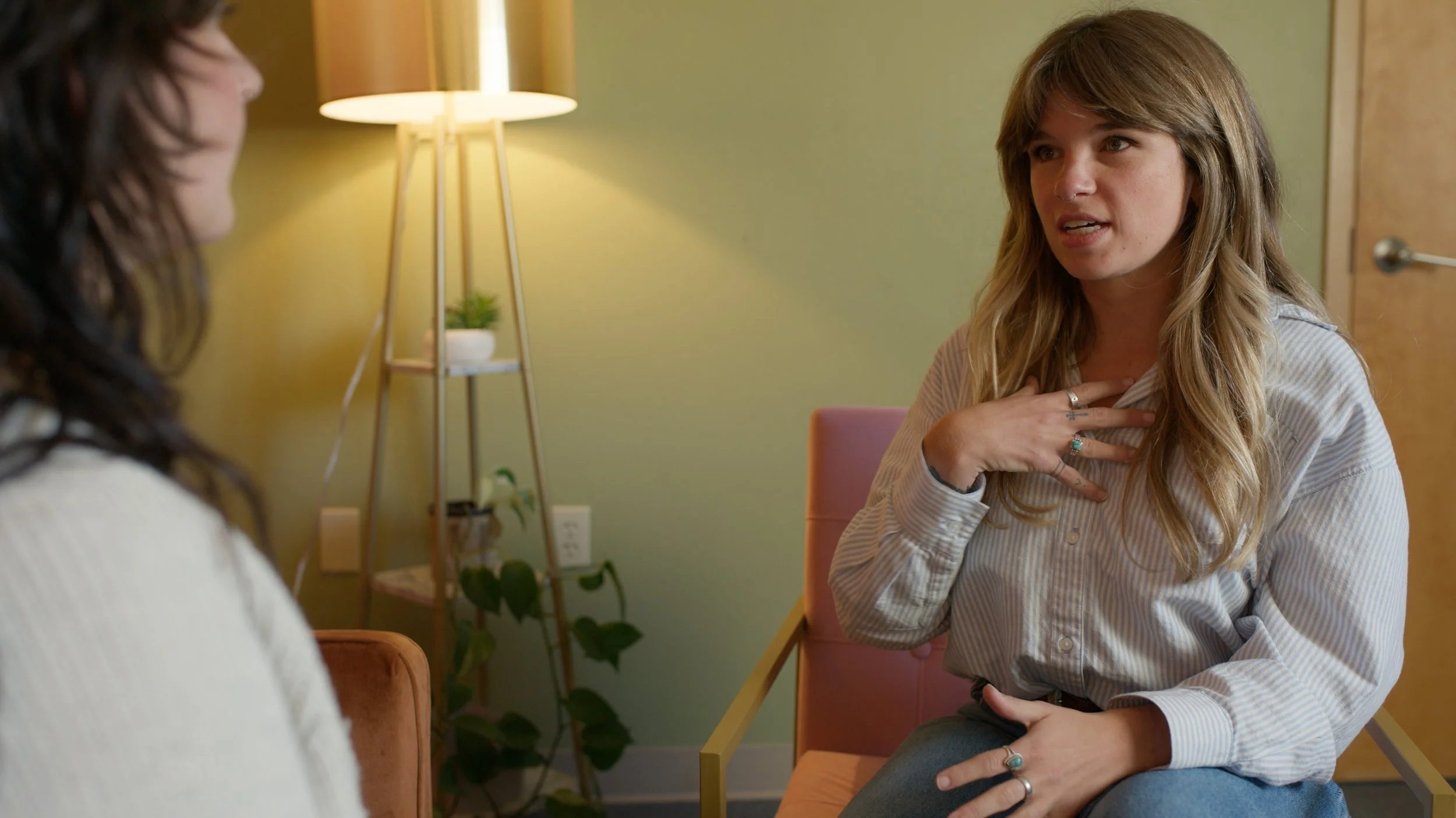 Two women engaging in conversation in a cozy, well-lit room with a yellow-green wall and a lamp in the background.