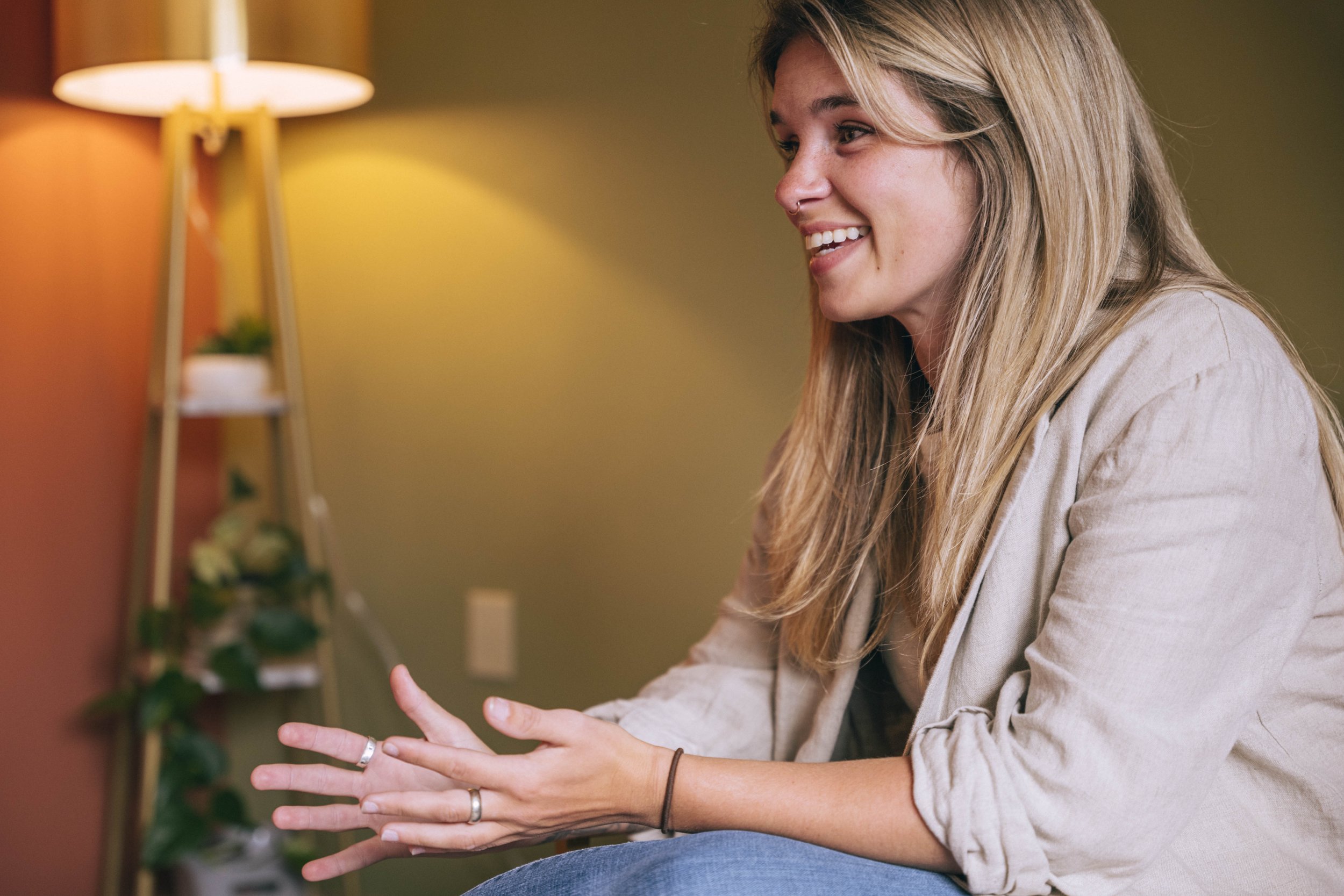 A woman with long blonde hair, smiling and speaking, wearing a beige blazer and which is rolled up at the sleeves, and sitting in a room with warm lighting and a plant in the background.