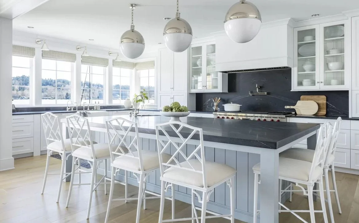 Bright kitchen with white cabinets, dark countertop island, large windows, and white chairs around the island.