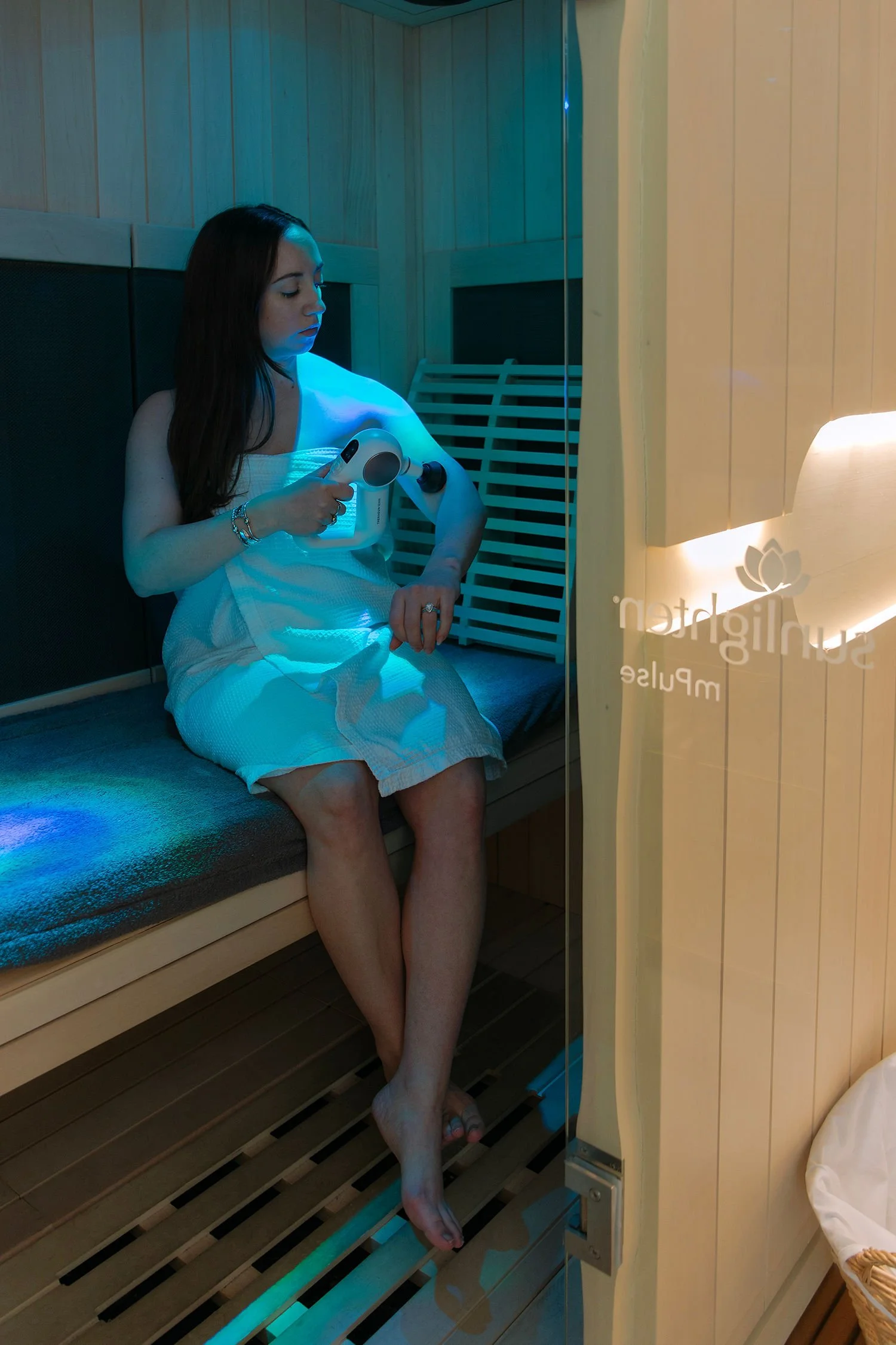Client using a massage tool during an infrared sauna session with chromotherapy lighting at Revival Wellness Studio