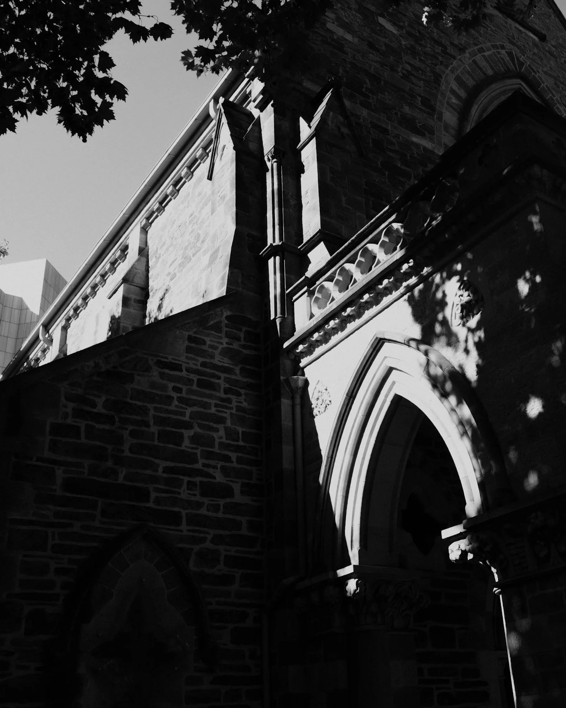 Black and white photo of a historic stone building with Gothic architectural details, including arched windows and decorative stonework, partially shaded by tree leaves.
