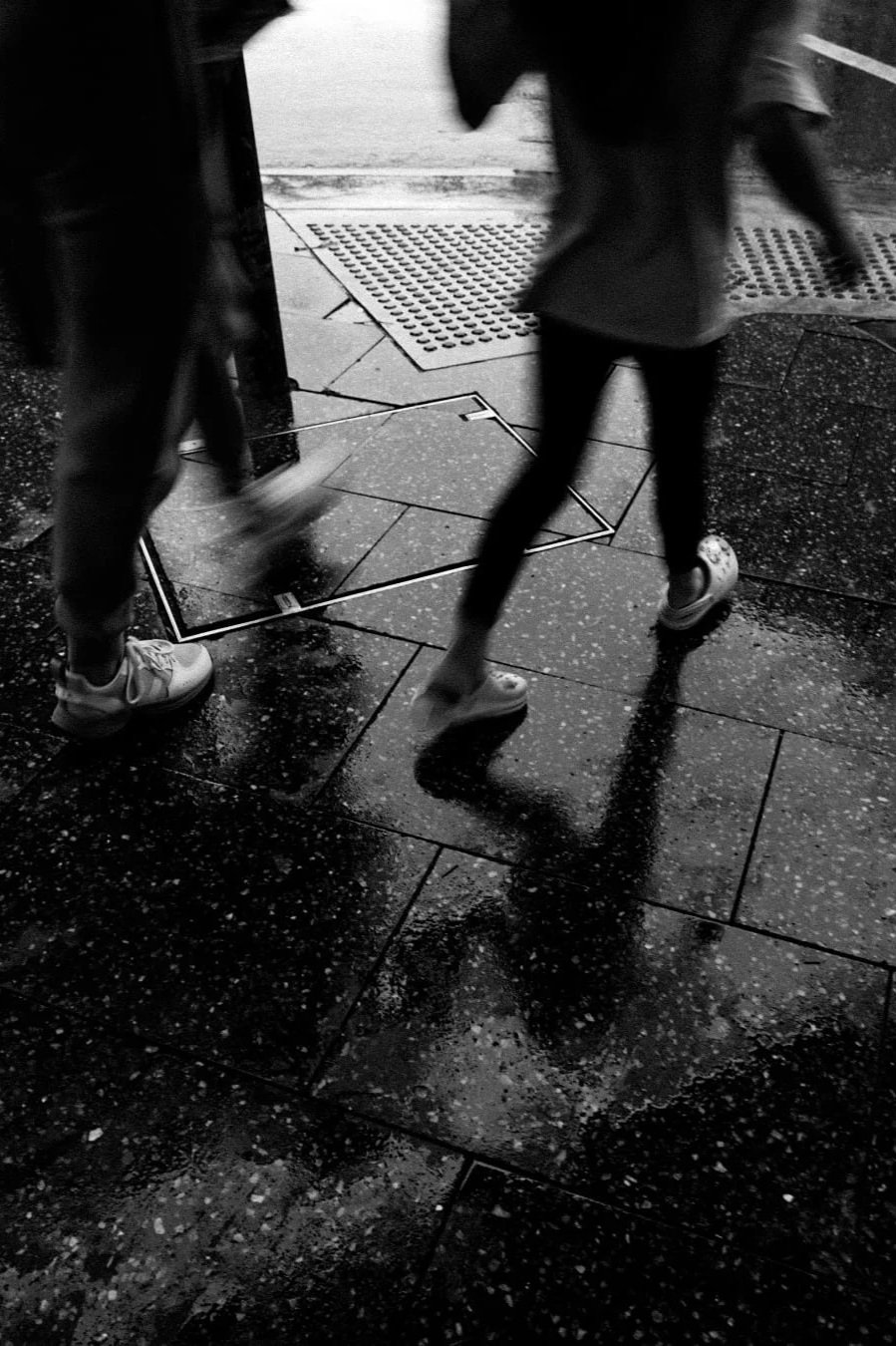 People walking on a wet city sidewalk at night, with reflections and shadows on the pavement.