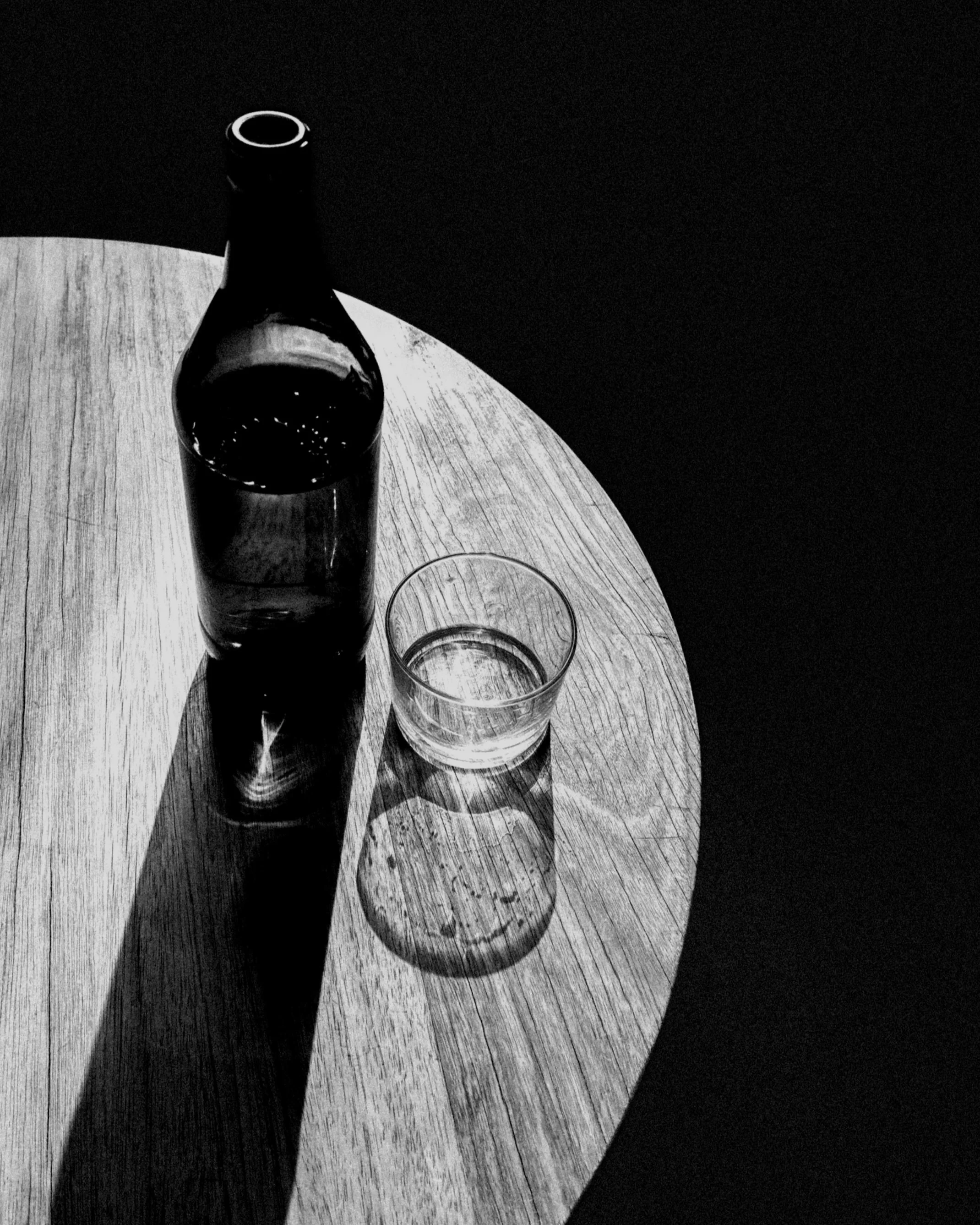A glass bottle and a small glass of water on a wooden `, with shadows cast on the surface in black and white.