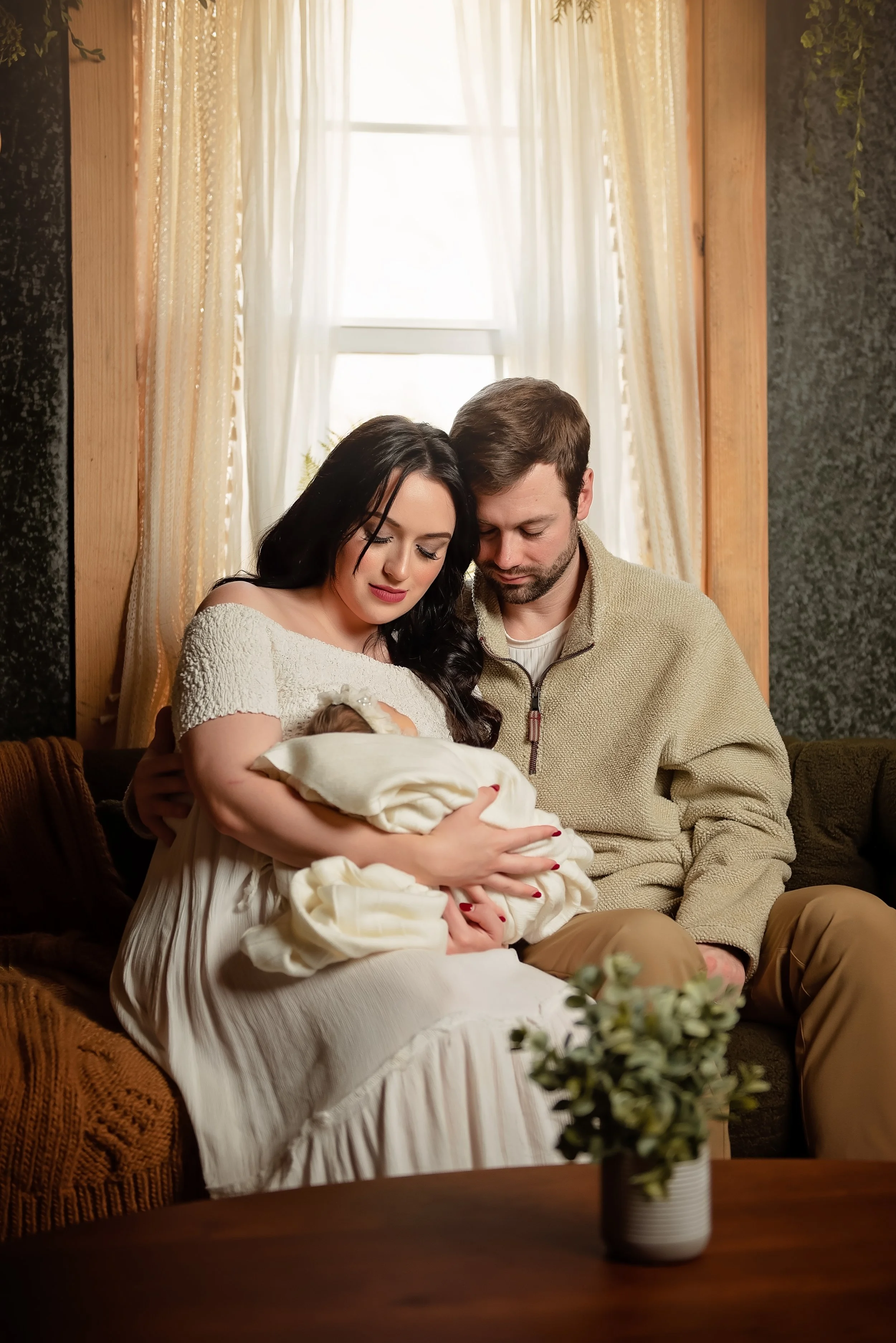 A family of three sitting on a sofa in a cozy room with sunlight streaming through a window behind them. The woman holds a newborn baby wrapped in a blanket, while the man sits closely beside them, all appearing to cherish a tender moment.