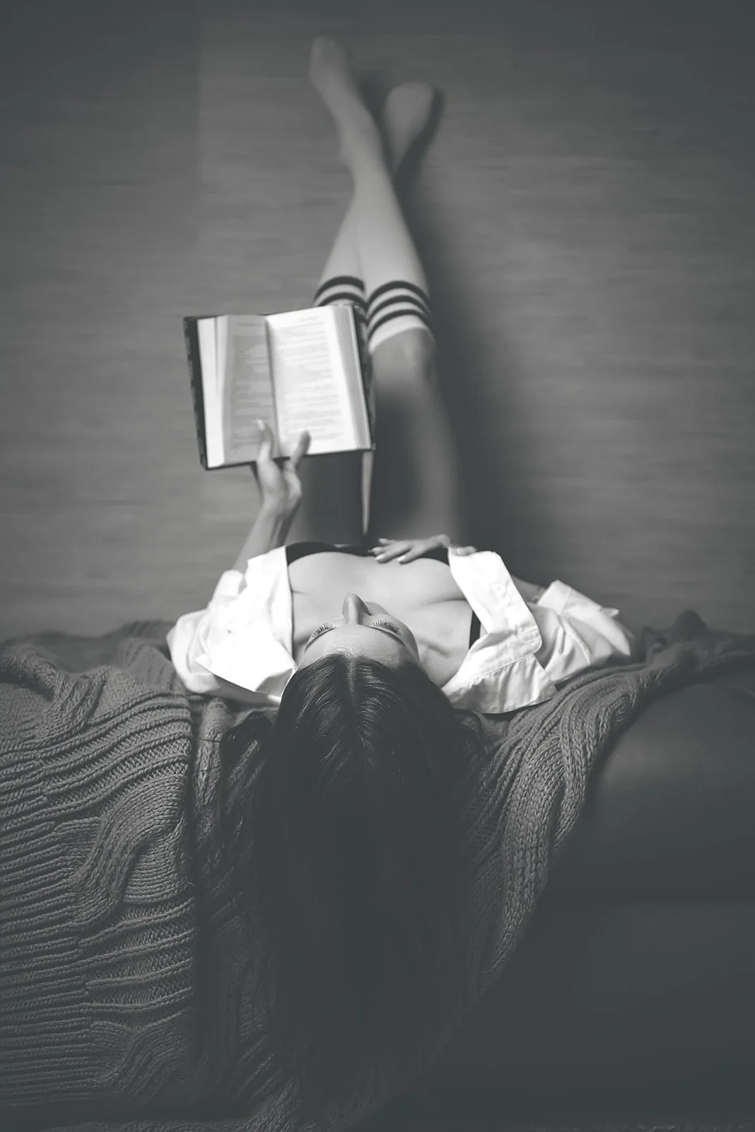 black and white photo of woman in unbottned shirt reading a book with her legs up the wall behind her. her hair flows down toward the floor