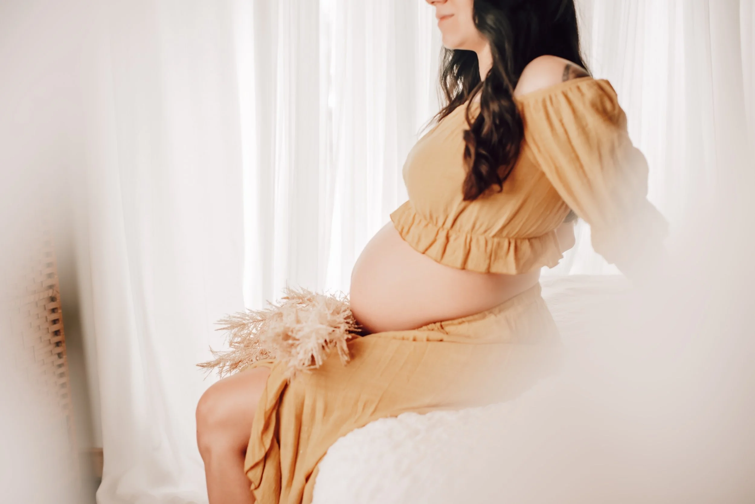 Pregnant woman sitting on a bed, wearing a yellow dress, with a soft-focus background of white curtains.