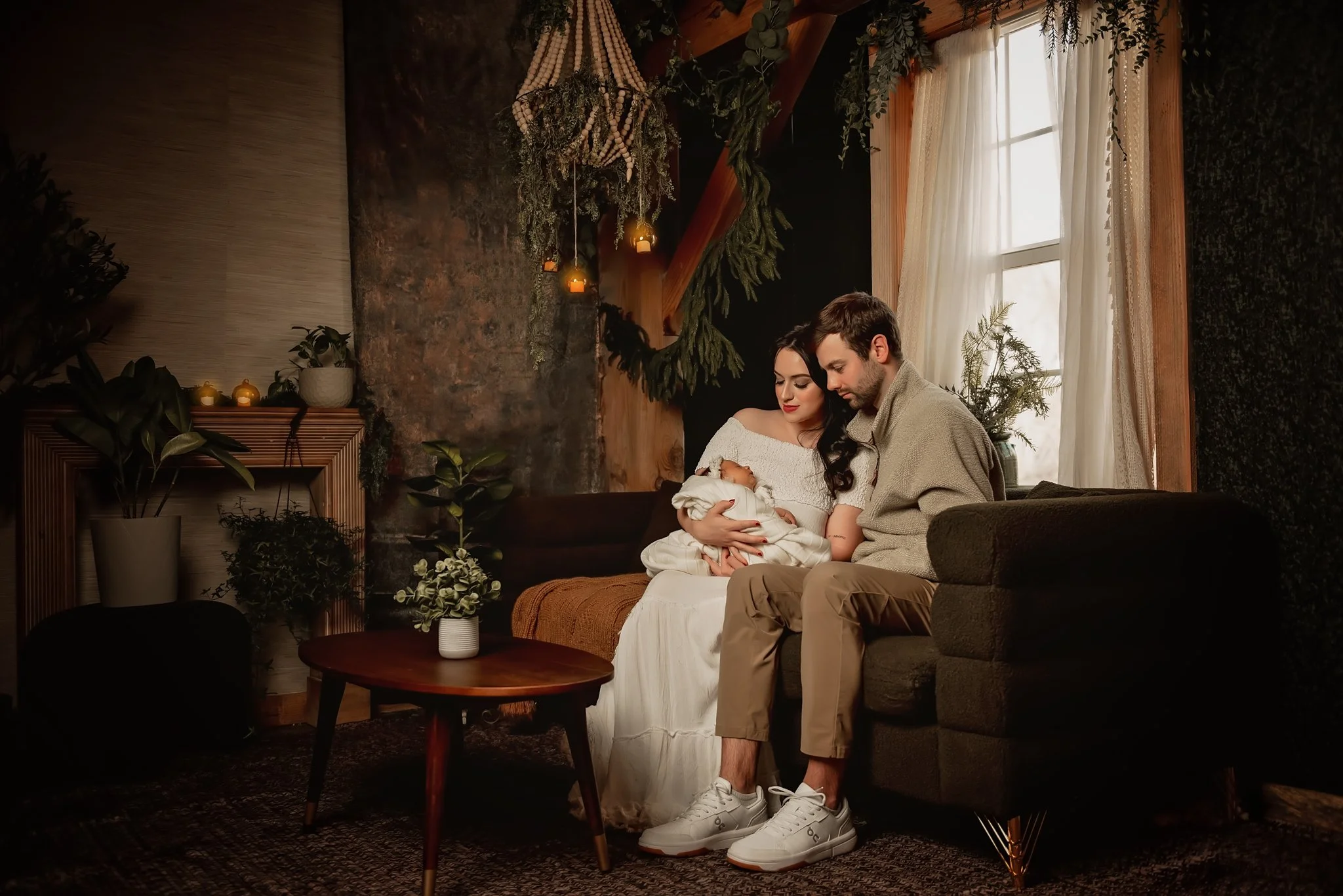 A family of three sitting on a couch in a cozy, warmly decorated room. The mother is holding a newborn baby wrapped in a blanket, and both parents are looking affectionately at the baby. The room has natural light coming through a window with light-colored curtains, and there are plants and candles adding to the warm ambiance.
