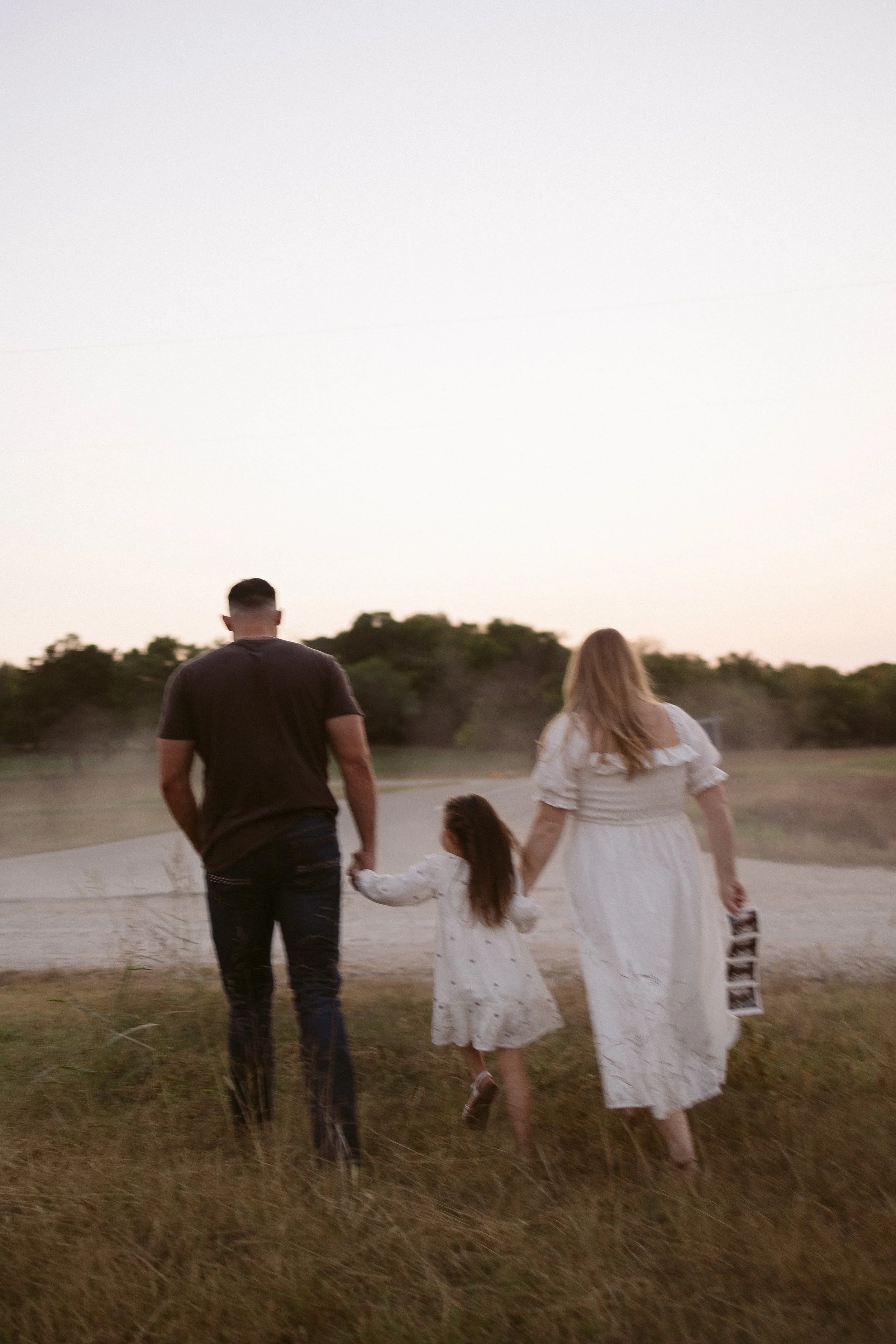 A family of three, consisting of a man, woman, and young girl, walking hand in hand on a grassy field during sunset, with trees in the background.
