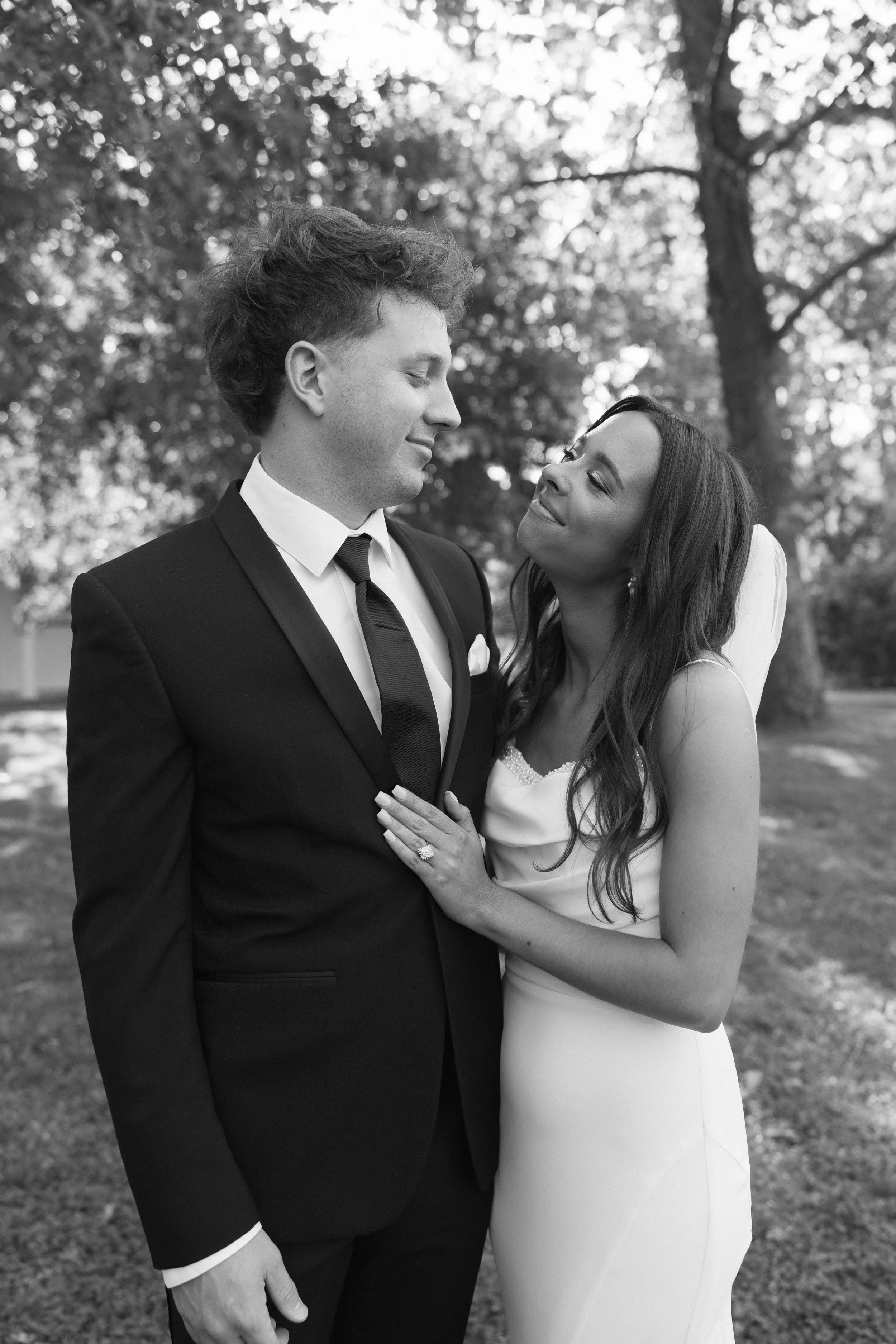 Black and white photo of a bride and groom in outdoor setting, gazing at each other lovingly, with trees in the background.