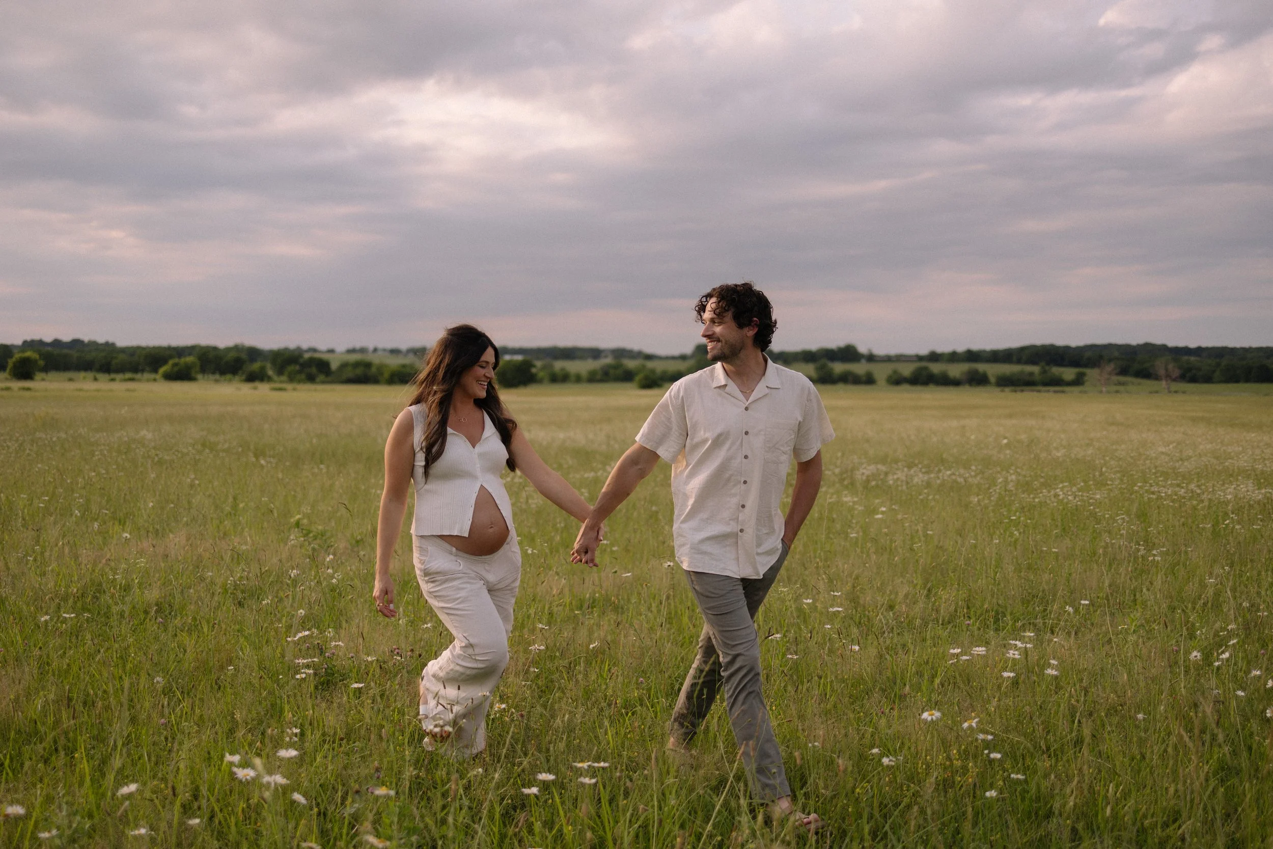 A pregnant woman and her partner holding hands and walking in a grassy field under a cloudy sky.