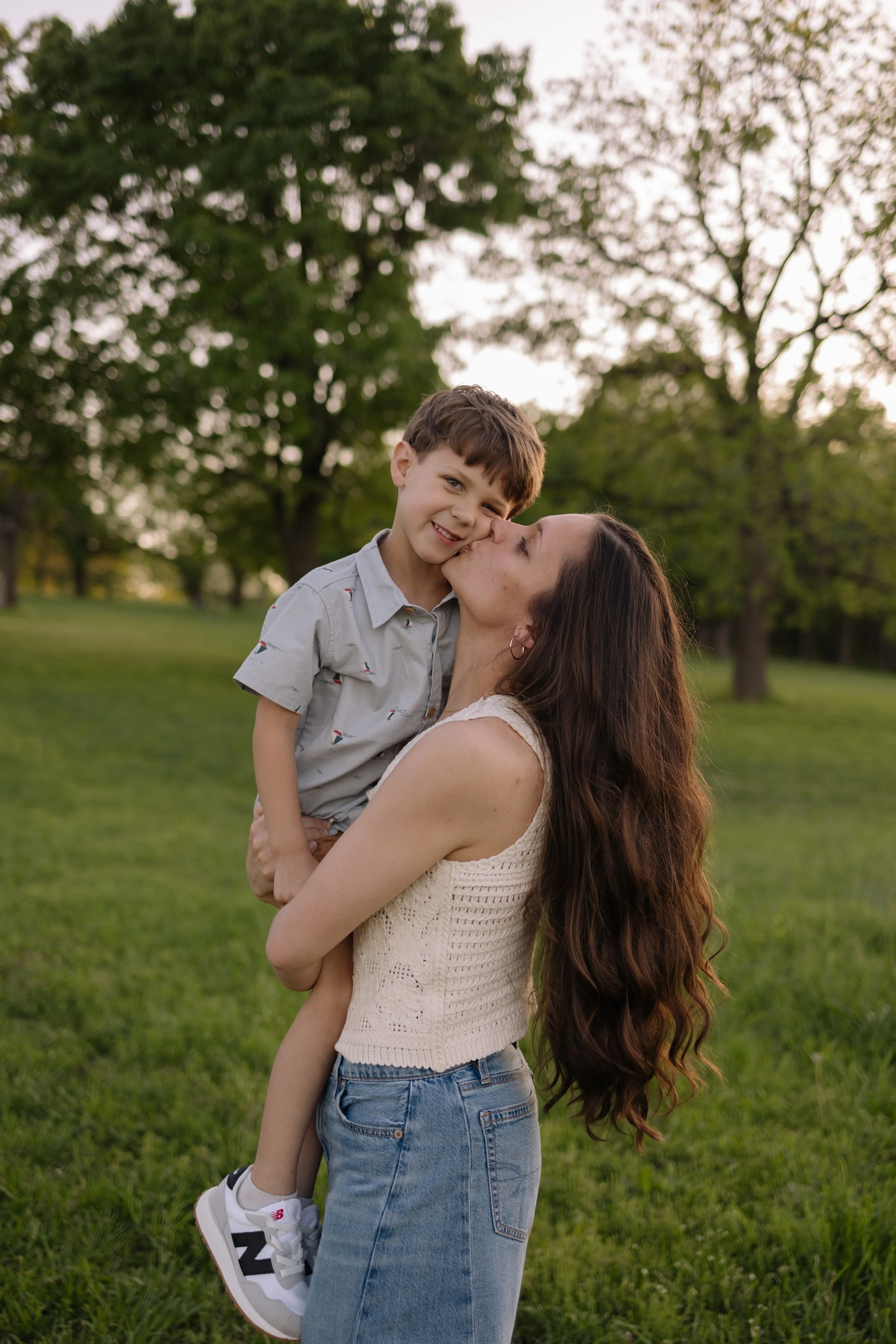 A woman with long, wavy brown hair lifting a young boy in her arms in a park with green grass and trees.