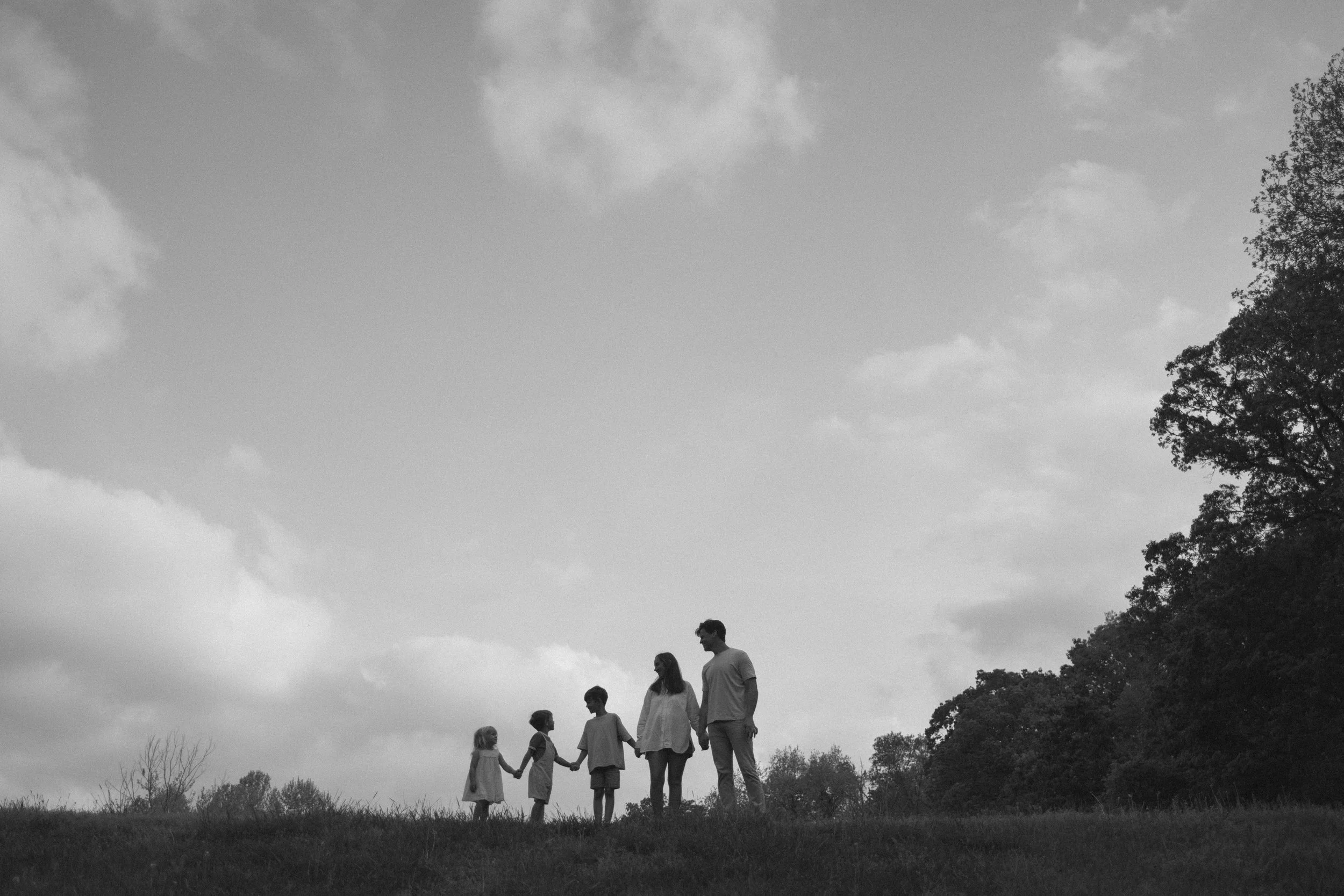 A black and white photo of a family of six standing in Bentonville Arkansas on grass in a park, holding hands, with trees in the background under a partly cloudy sky.