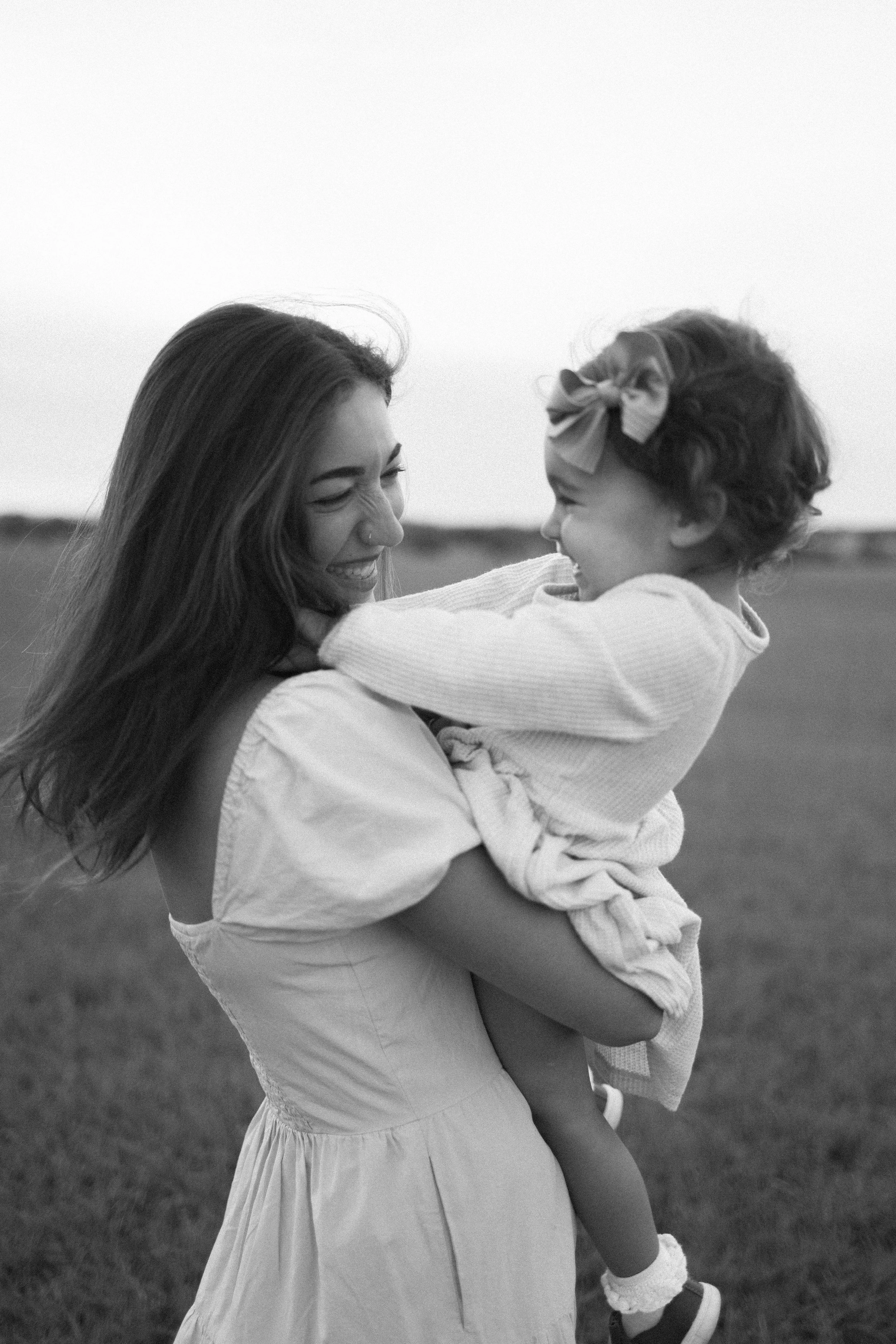 A woman is holding a young girl in an open outdoor field. They are smiling and appear to be playing or sharing a joyful moment together.
