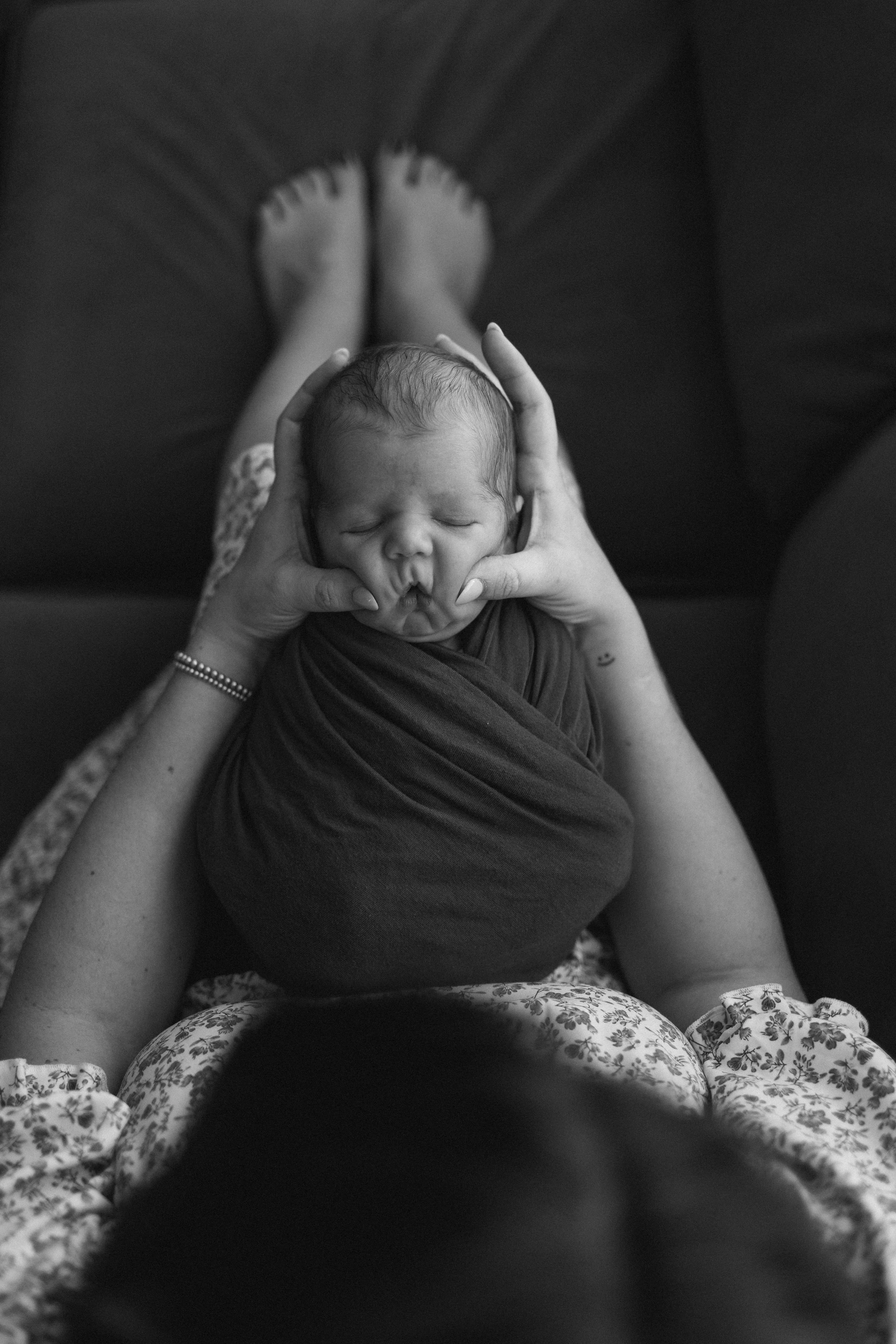 Black and white photo of a newborn baby wrapped in a blanket, being held gently by an adult, with the adult lying on a couch and the baby's face relaxed with closed eyes.