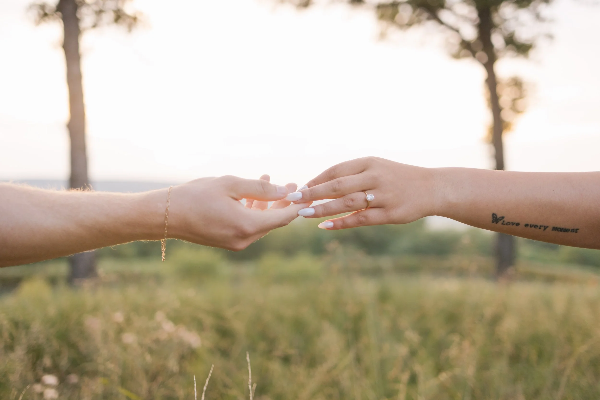 Two hands reaching towards each other, one with an engagement ring, in an outdoor setting with trees and a sunset background.