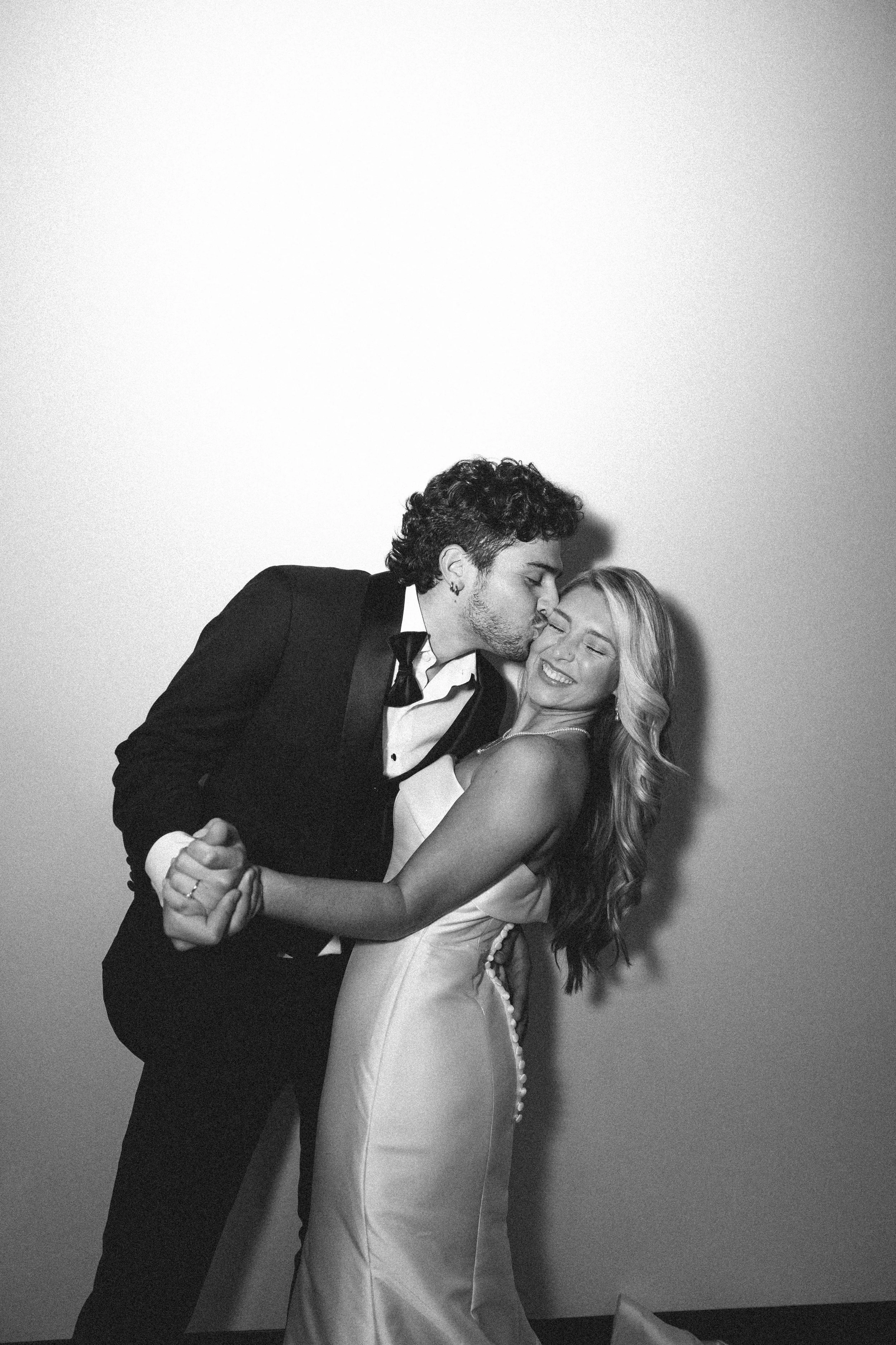 A black and white photo of a couple dancing closely; the man is in a tuxedo and is kissing the woman's forehead, she is wearing a satin dress and is smiling.