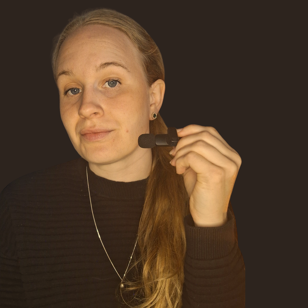 En kvinna med långt, rött hår som håller i ett headset med mikrofon framför ett mörkt bakgrund.