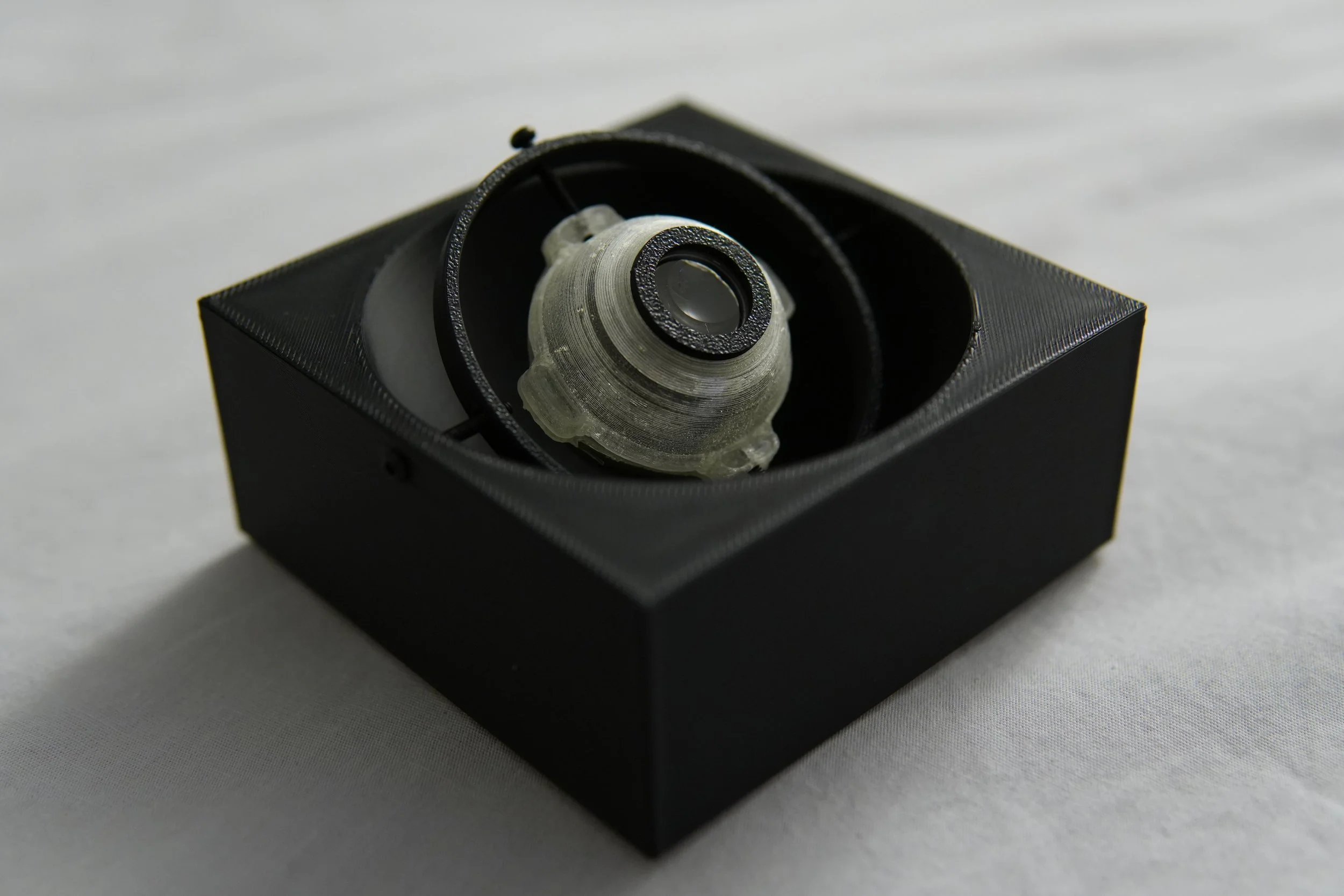Close-up of a small, intricate mechanical component inside a black box with a textured surface, placed on a white surface.